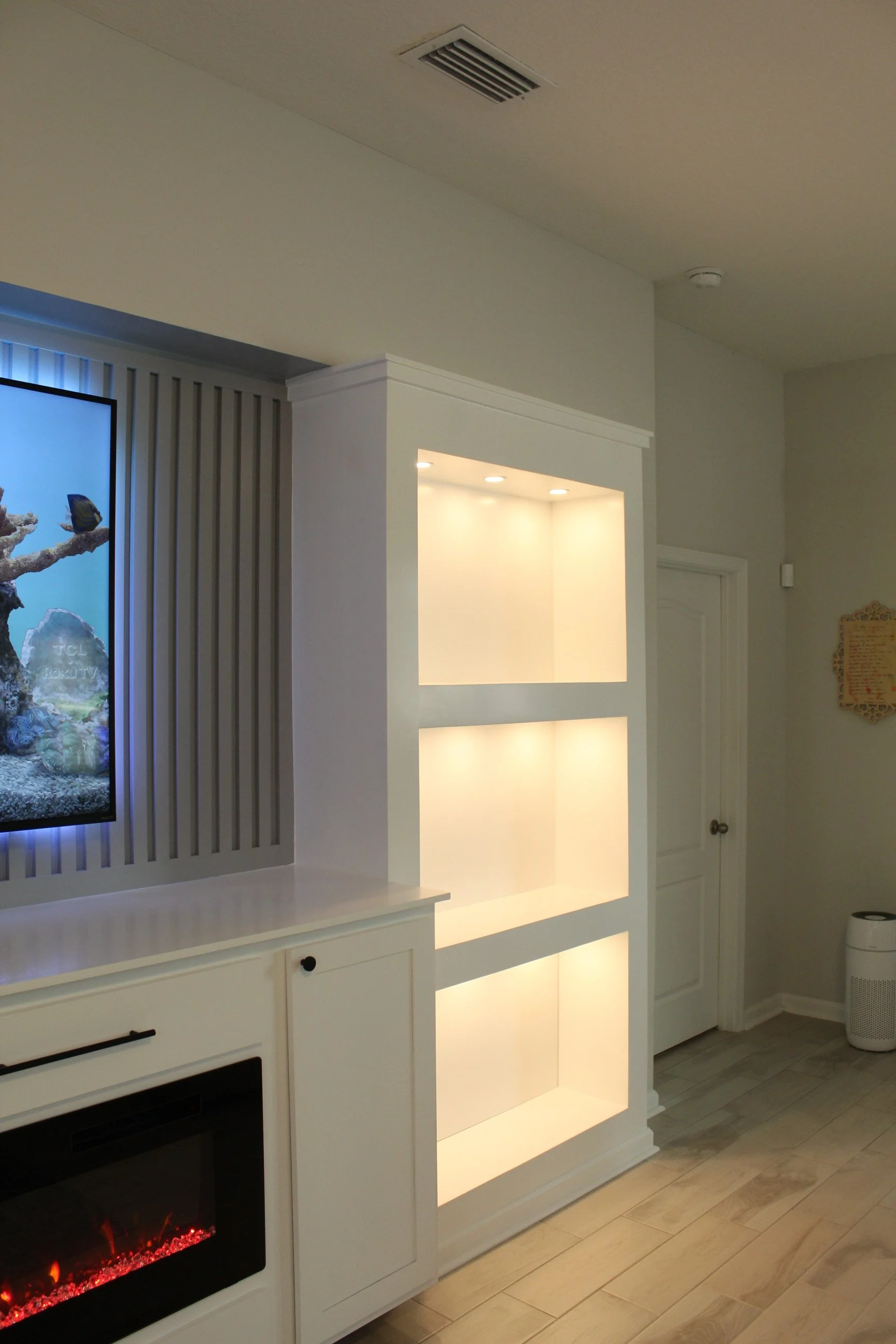 Interior of a room featuring a white cabinet with open shelves illuminated by built-in lights, an electric fireplace with a glowing ember effect, and a wall-mounted television on the left side. The floor is made of light-colored wood, and there is a closed door and a wall with a decorative frame on the right side.