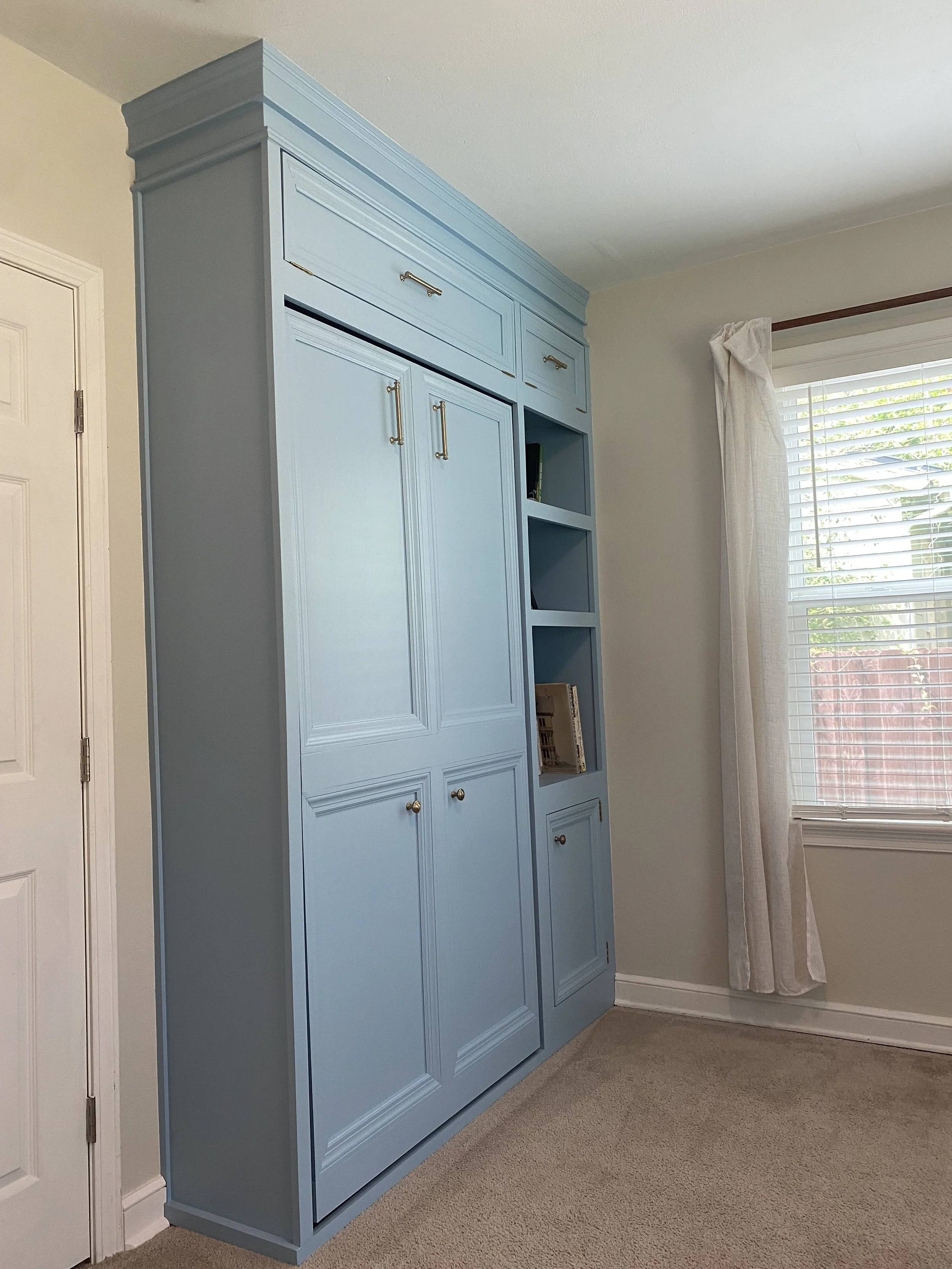 Blue built-in bookcase with gold handles in a room with white walls and a window with white blinds and curtains.