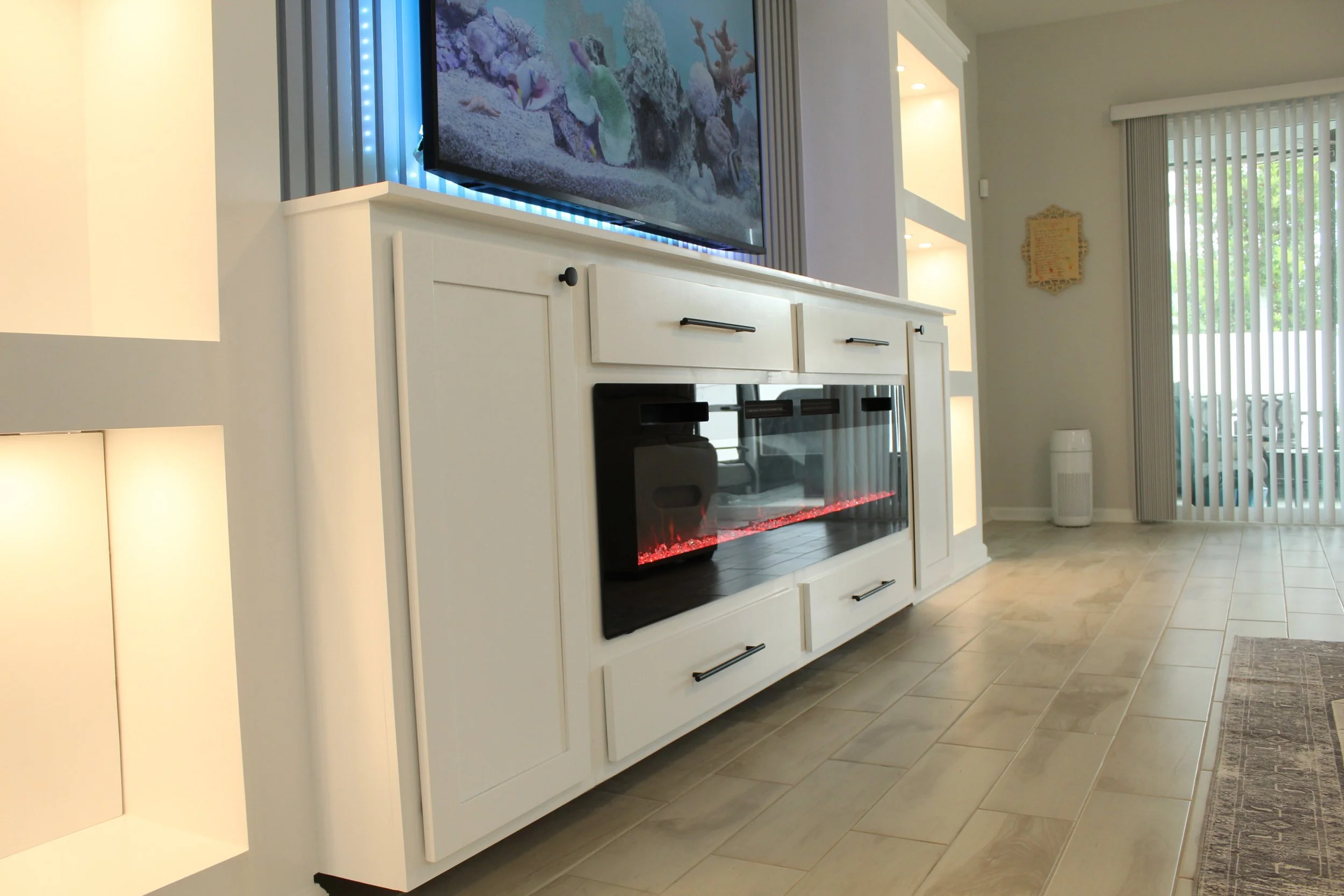 Living room with a white entertainment center, a large flat-screen TV displaying a coral reef scene, and an electric fireplace below the TV. To the right, there's a window with vertical blinds, a framed decoration on the wall, and a white air purifier on the floor.