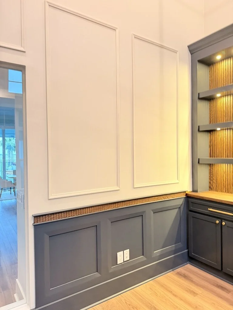 Interior corner of a room with light-colored wall panels and dark gray wainscoting. Built-in shelves with lighting and a wooden countertop are visible.