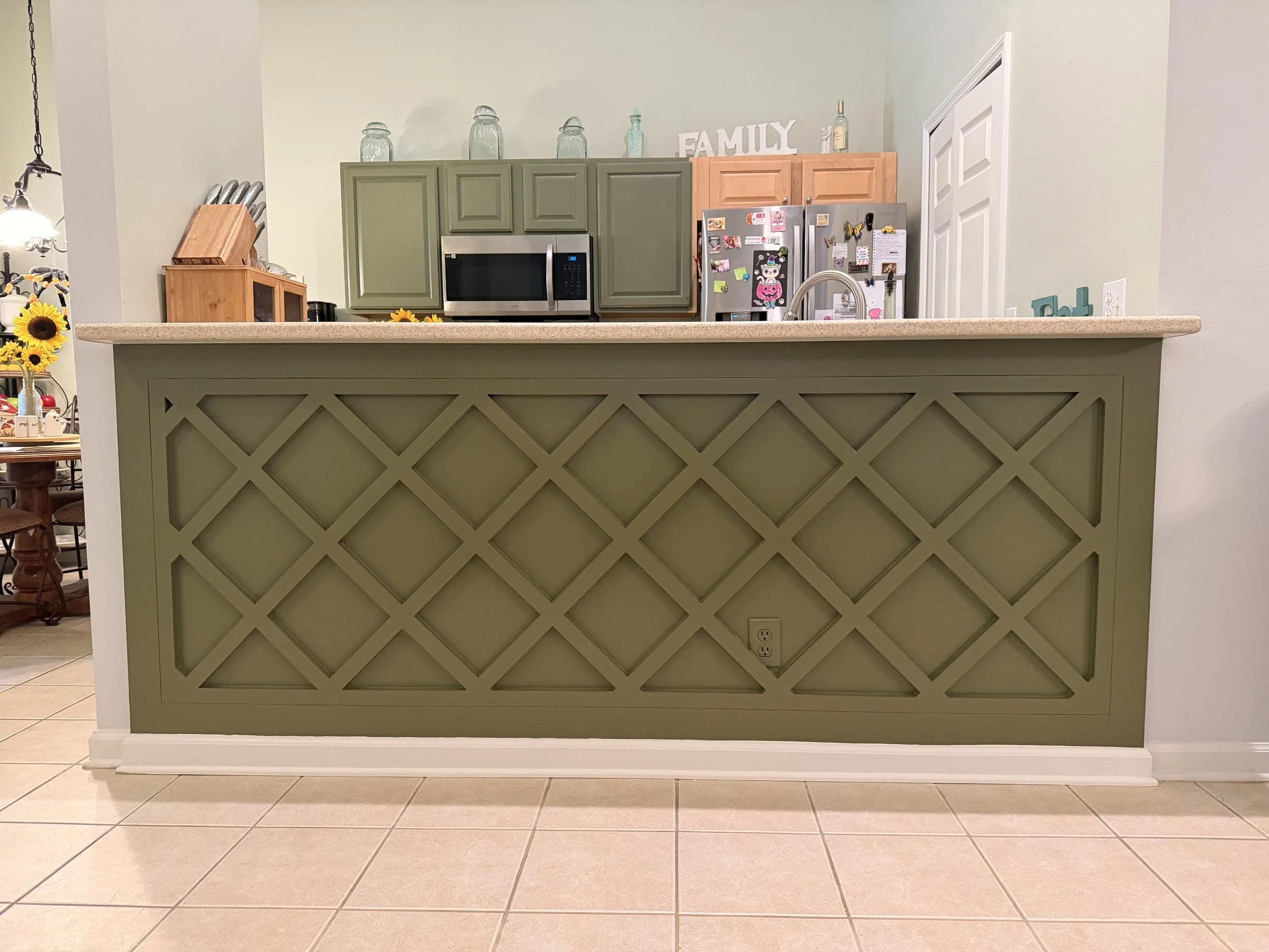 A kitchen kitchen with green cabinets, a microwave, and a refrigerator covered with magnets and notes. There is a countertop with a sink. Behind the countertop, decorative glass jars and a sign that says 'FAMILY' are on top of the cabinets. Part of a dining area with a table and chairs is visible to the left.