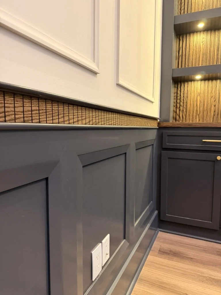 Close-up of a kitchen wall with dark gray wainscoting, two electrical outlets, and part of a wooden floor and cabinetry with wall-mounted shelves