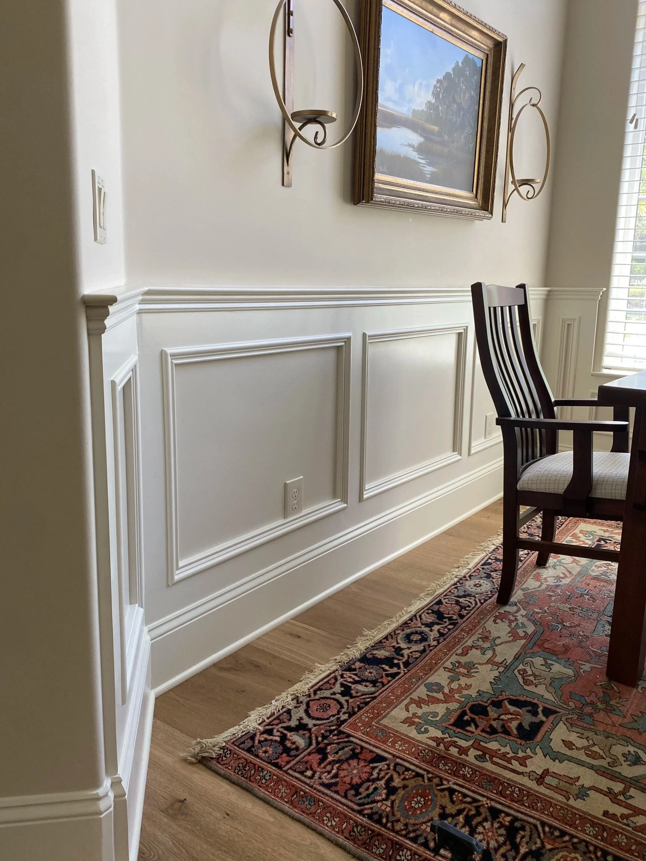 A dining room or living space with white wainscoting and trim on the wall, a framed landscape painting of a river, trees, and sky, two wall-mounted candle holders, a wooden chair with a cushioned seat, a patterned area rug with fringes, and part of a window with white blinds.