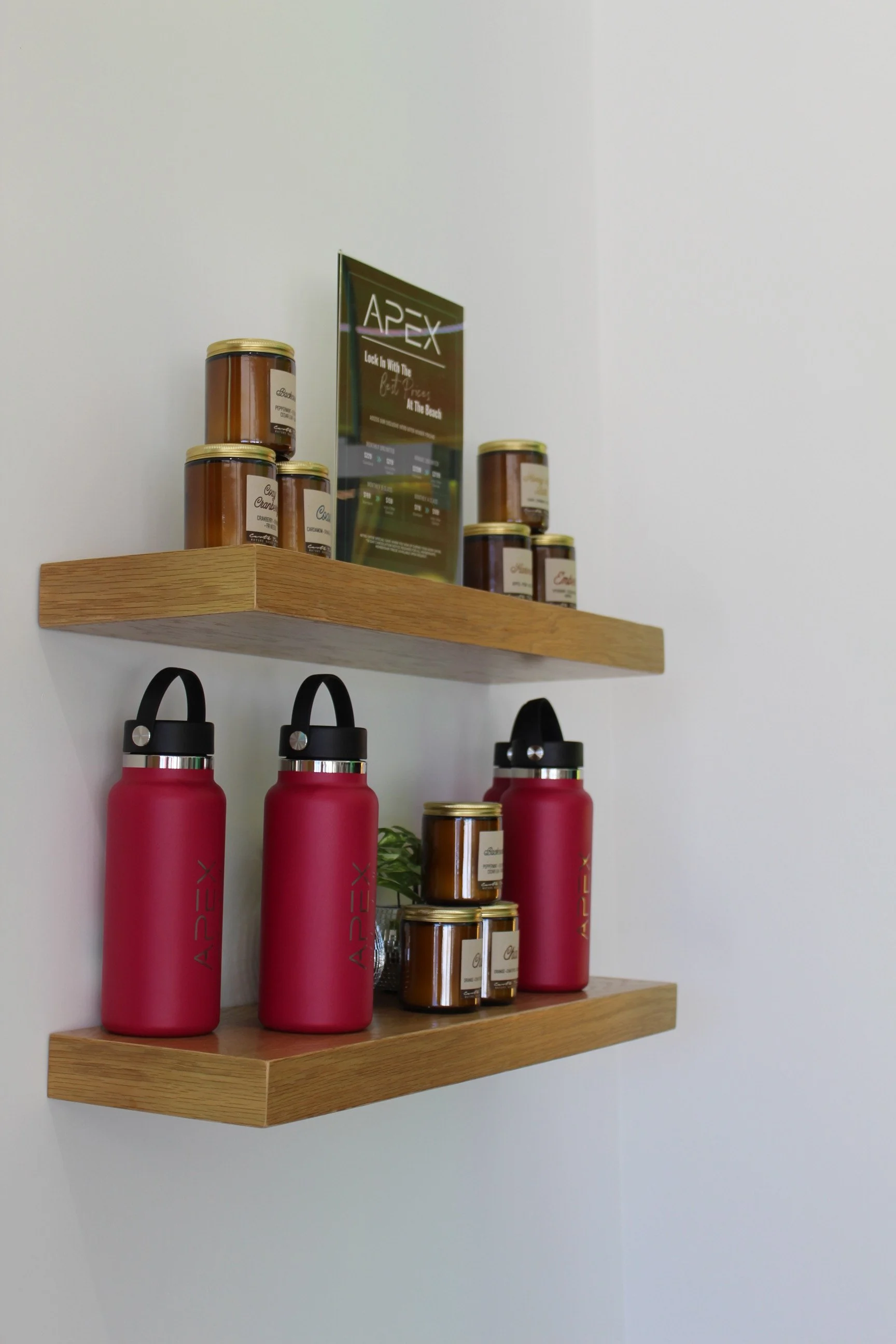Two wooden shelves on a white wall display bottles of pink reusable water bottles, jars of coconut caramel spread, and a framed Apex advertisement sign.