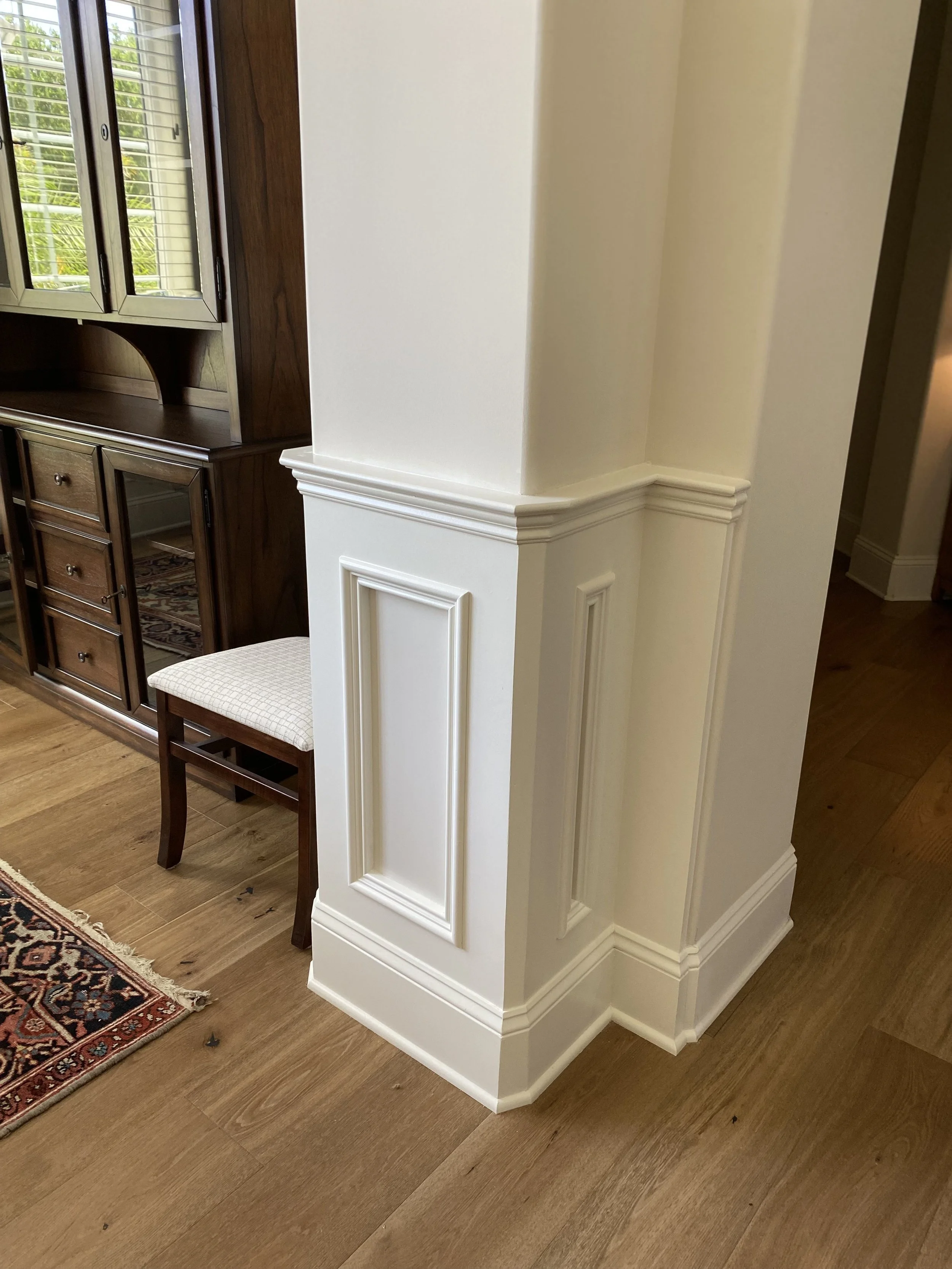 Interior view of a room corner with a white wainscoting wall trim and a section of a dark wooden cabinet with glass doors, next to a wooden chair with a cushioned seat, on a hardwood floor with a patterned rug nearby.