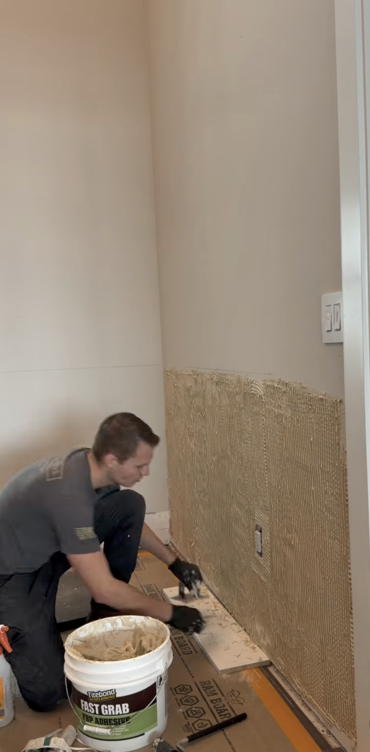 A man kneeling on the floor applying adhesive to a wall with a notched trowel, next to a white bucket of tile adhesive, preparing to install tile. The wall has a section with a textured backing material, and there is a power outlet on the wall.