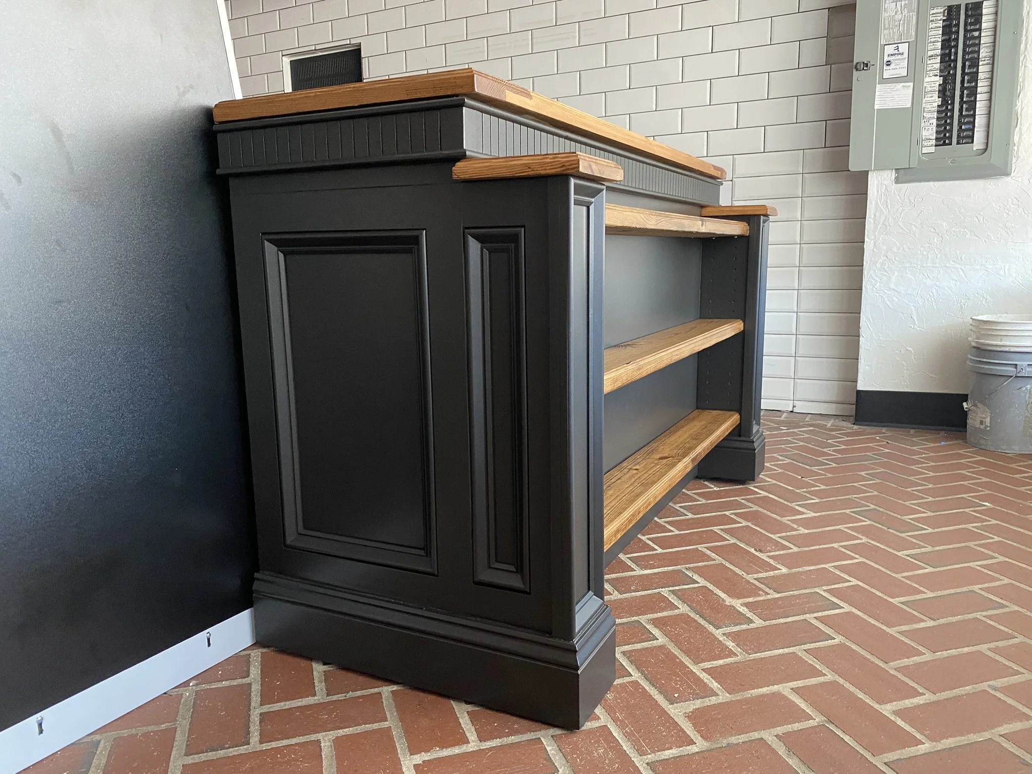 Empty black and natural wood display case with shelves, placed on a brick floor near a wall with a gray electrical panel.