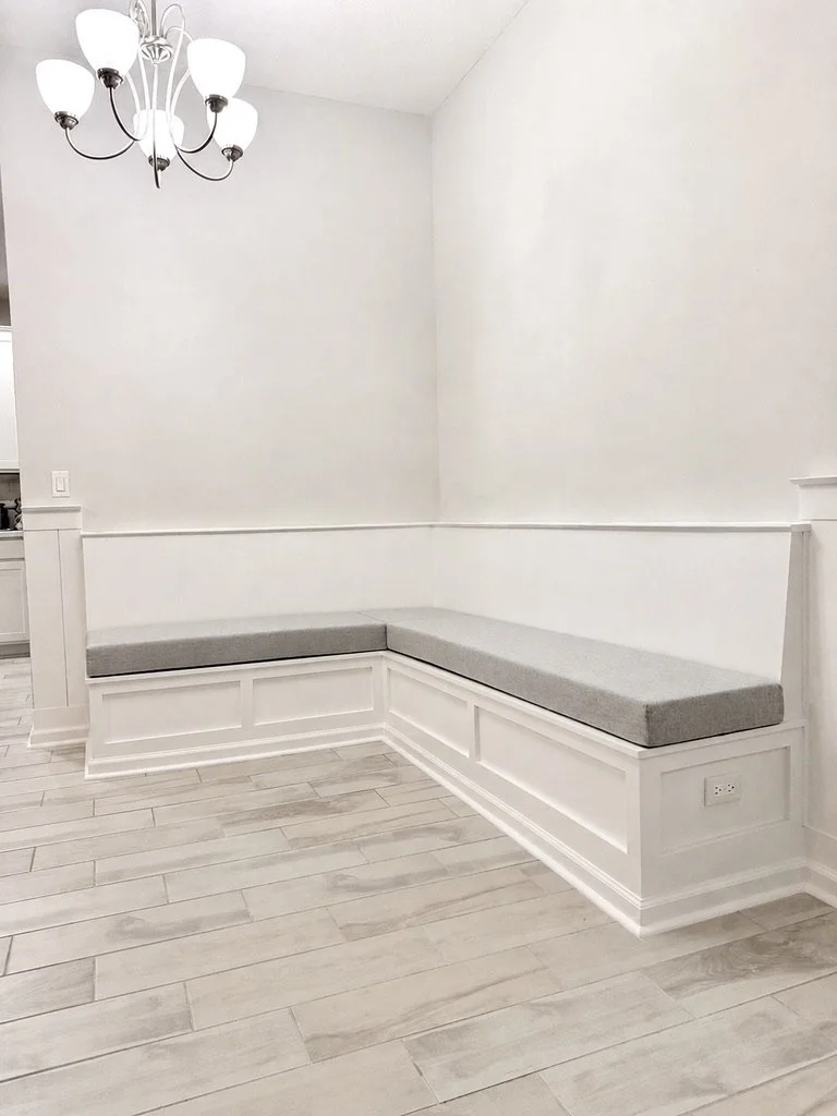 White corner built-in bench with gray cushions in a room with white walls, light wood flooring, a chandelier, and electrical outlet.