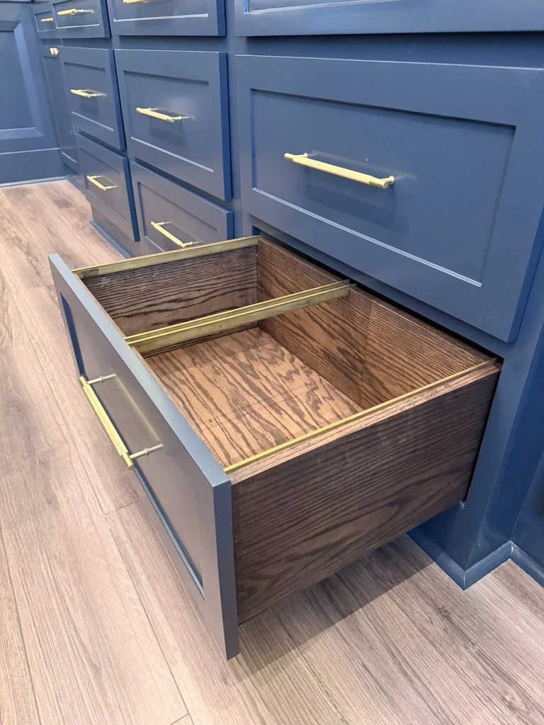 An open kitchen drawer with a wooden interior and gold-colored handles, part of blue cabinetry with similar drawers and gold handles, on a wooden floor.