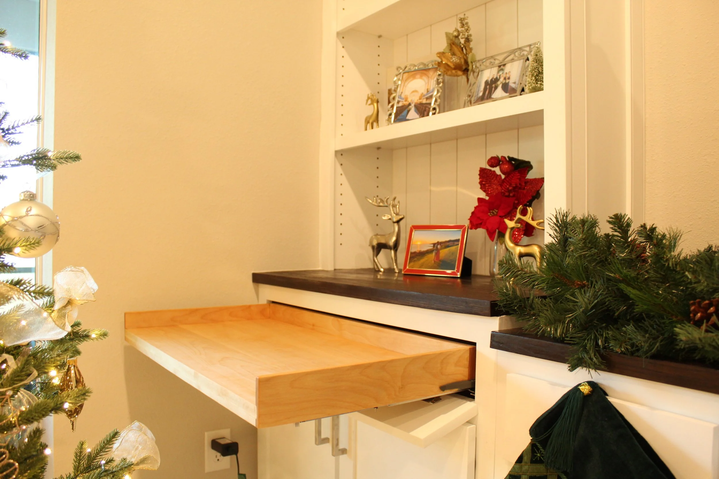 Decorative Christmas shelf with framed photos, gold reindeer, and poinsettia plant, near a decorated Christmas tree with ornaments and ribbons in a cozy home interior.