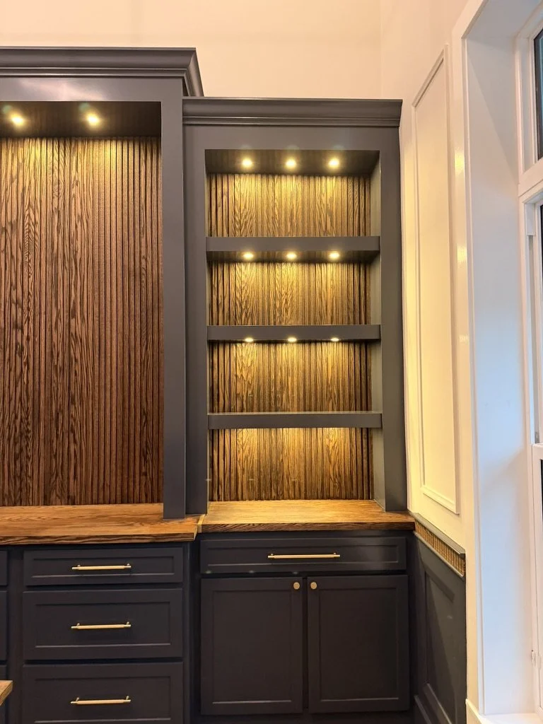 A built-in cabinet with open shelves featuring vertical wooden paneling and warm lighting. The lower section has a dark finish with drawers and cabinet doors, while the upper section includes three open shelves illuminated from above.