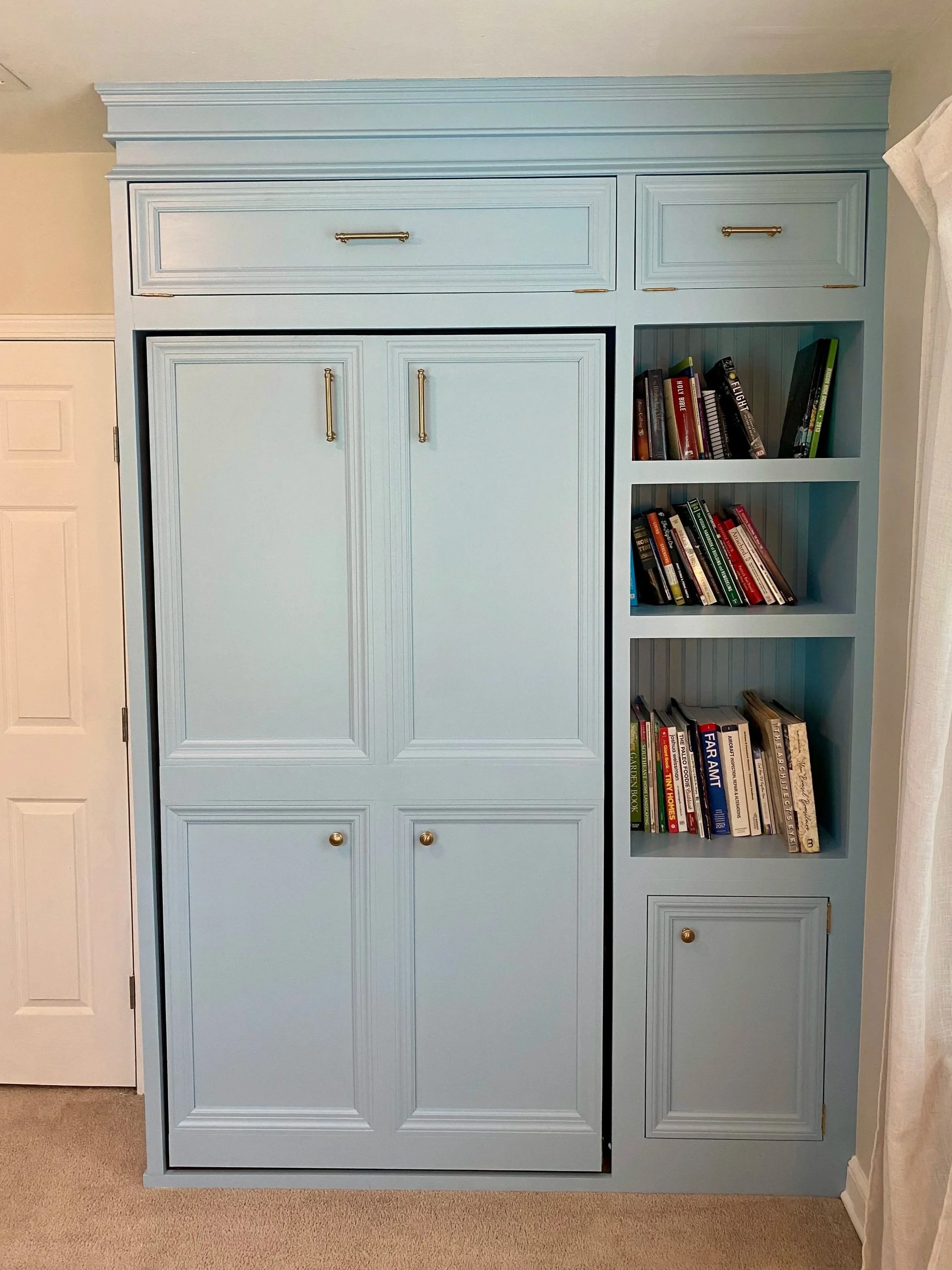 Light blue built-in bookshelf with closed cabinets on the left and open shelves on the right, filled with books, situated in a room with beige carpet and a white door.