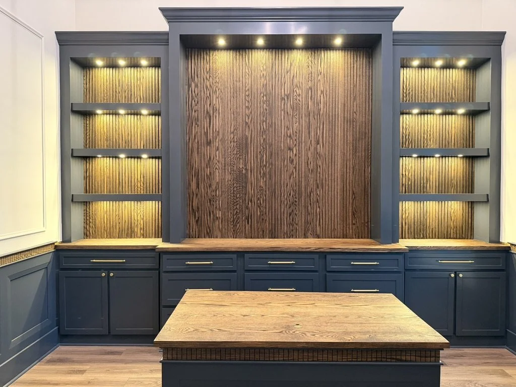 A built-in wall unit with open shelves and cabinets, painted dark blue, with a wooden backdrop and a matching wooden table in front, lit by recessed lights.