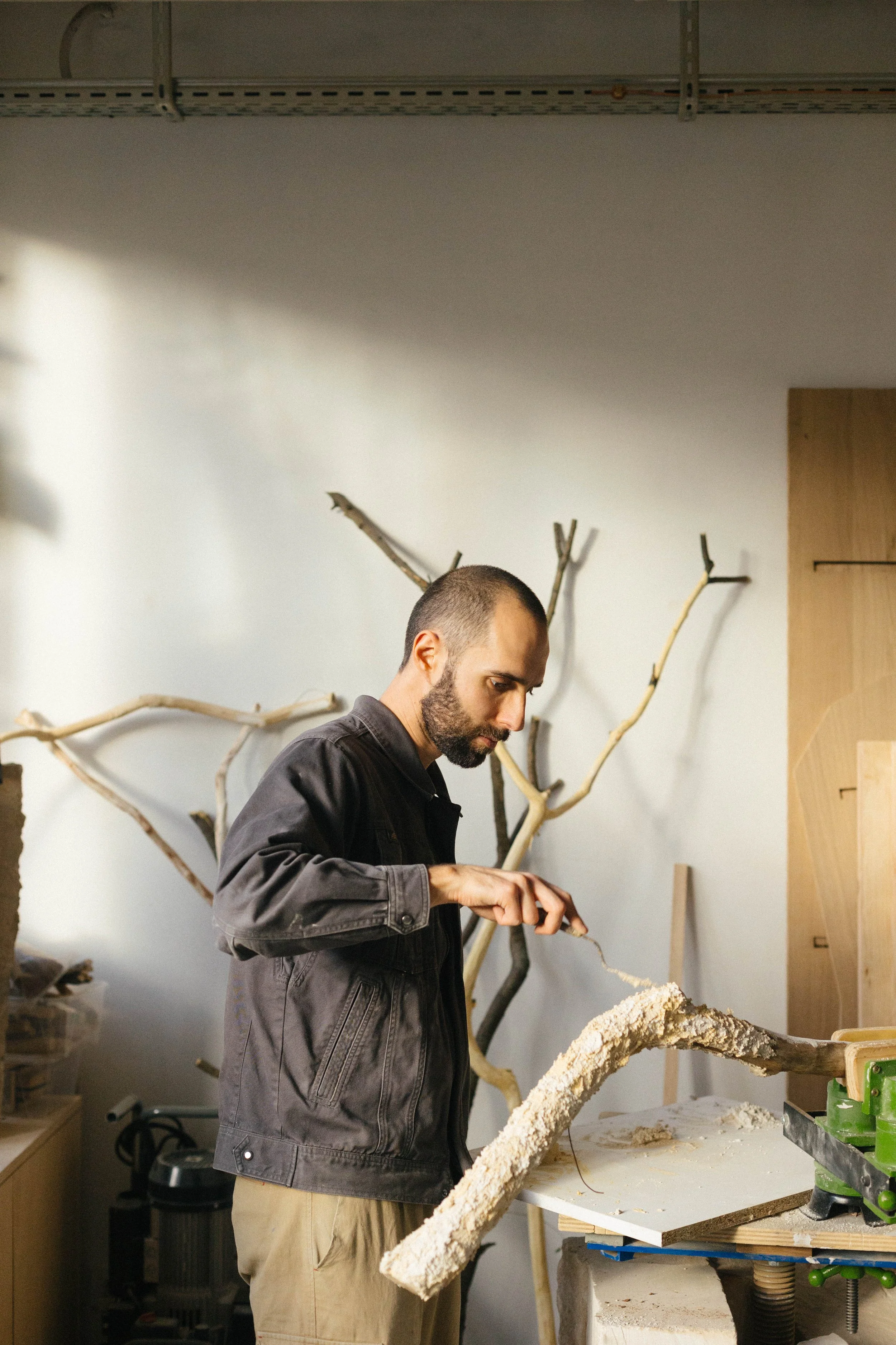 Un homme travaille dans un atelier, il manipule un objet en bois ou en matière similaire, avec des branches d'arbres ou bois d'œuvre en arrière-plan.