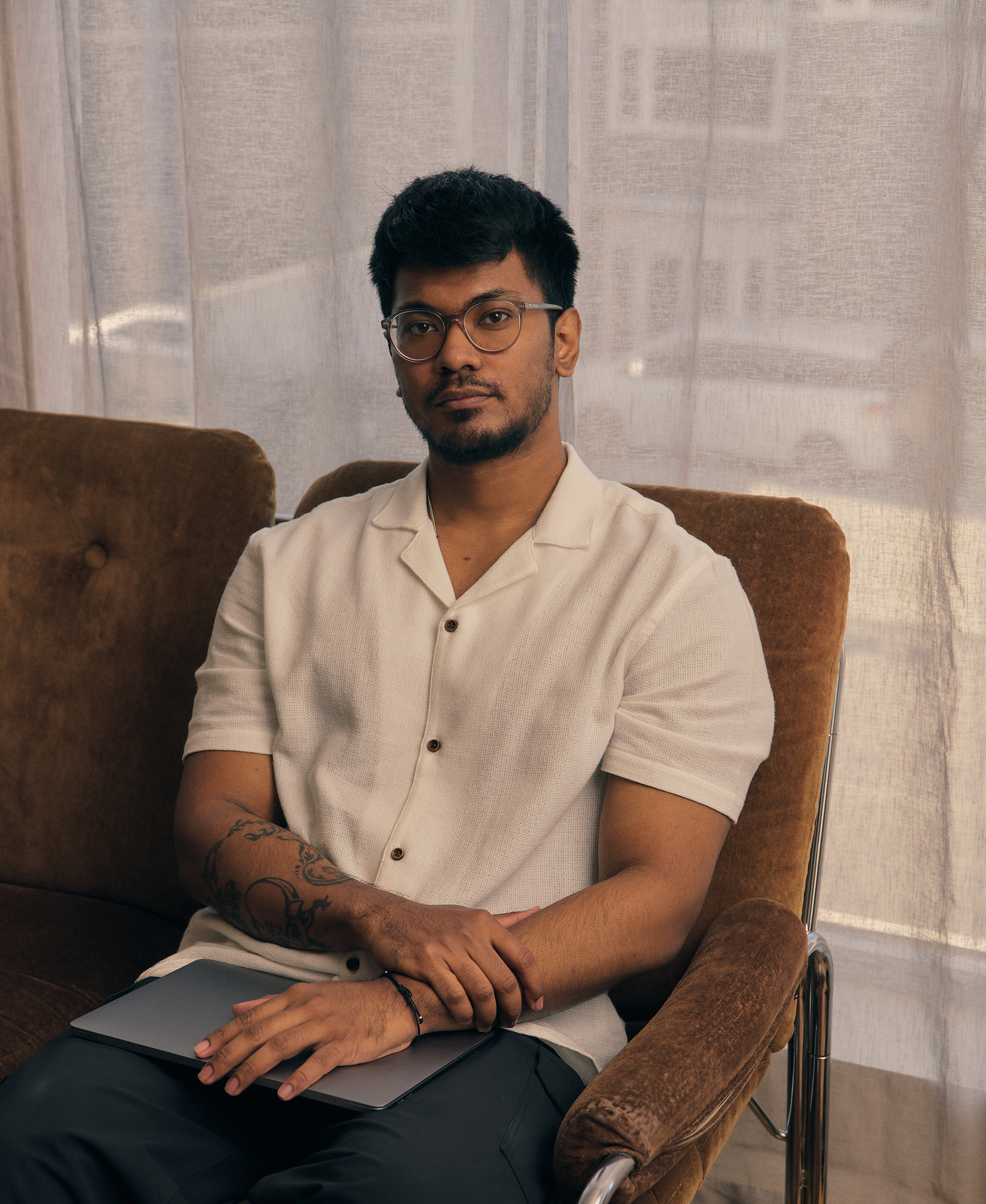 A young man sitting on a vintage brown armchair with black leather armrest and a wheel. He is wearing a beige short-sleeved button-up shirt and glasses. His left arm has a tattoo with a black design. A closed laptop is on his lap. Behind him, there is a translucent curtain with a sheer fabric that allows some natural light through, creating a soft ambiance.