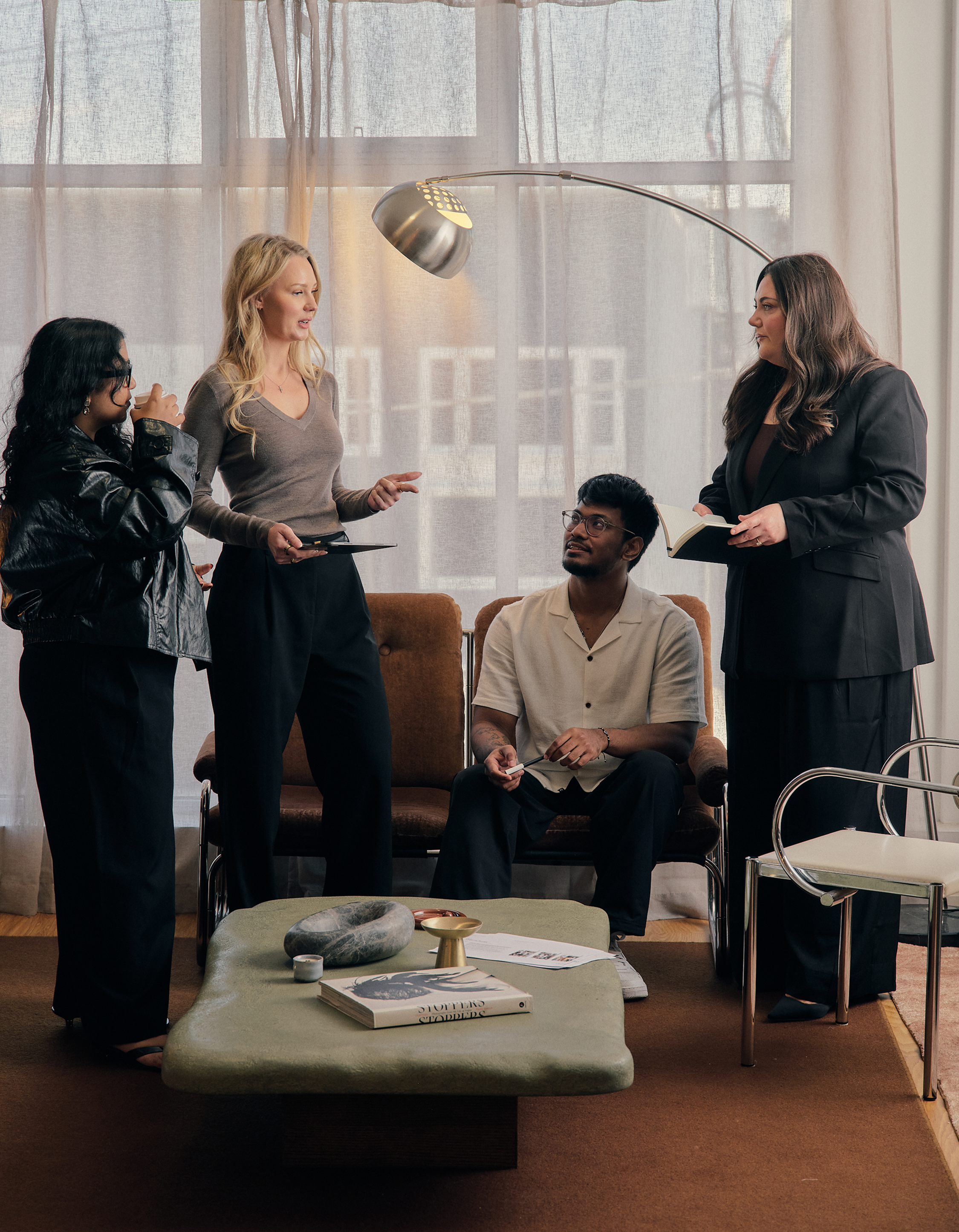 Four people in a living room having a discussion, with two women standing and talking, one man sitting on a chair, and another woman standing with a book. There is a coffee table with magazines and decorative objects in the foreground, and large windows with curtains in the background.
