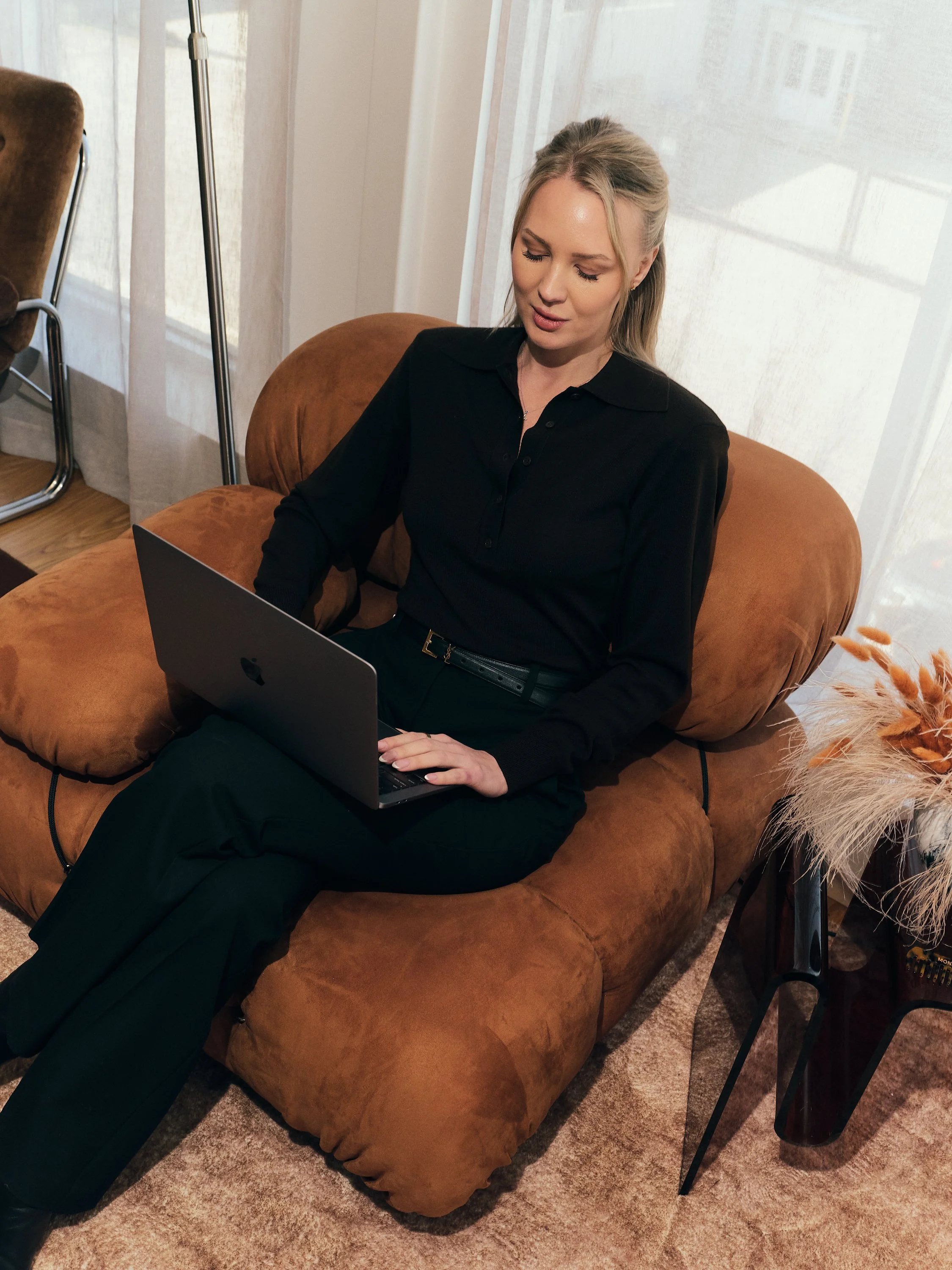 A woman with blonde hair in a half-up style, wearing a black long-sleeve shirt and black pants, sits on a brown velvet sofa working on a silver laptop in a cozy room with a beige carpet, a window with sheer curtains, a decorative vase with dried flowers, and a black side table.