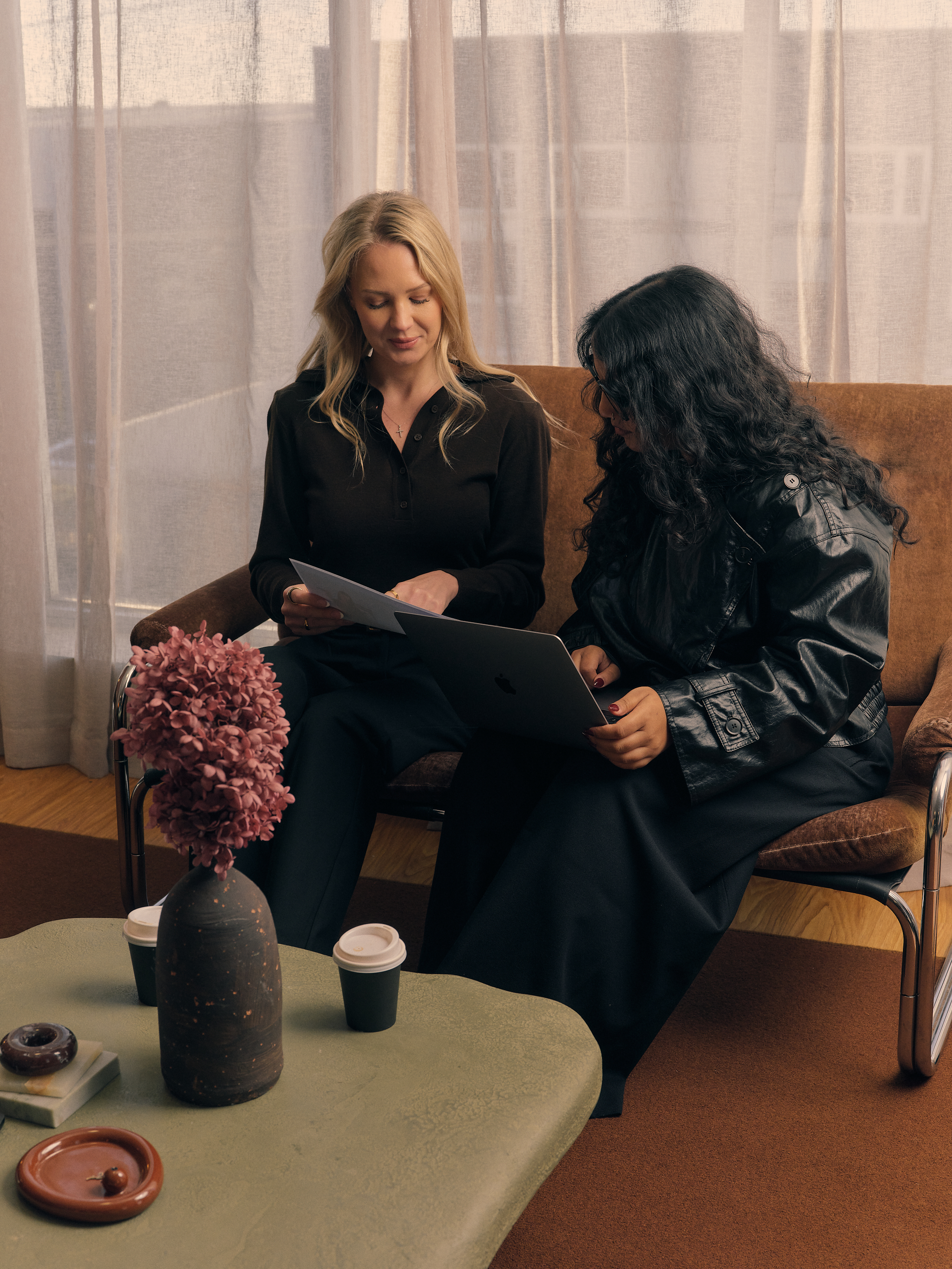 Two women sitting on a brown sofa in a living room, looking at a tablet together. One has blonde hair and the other has curly dark hair and wears glasses. A flower arrangement, cups of coffee, and small desserts are on a table in the foreground.