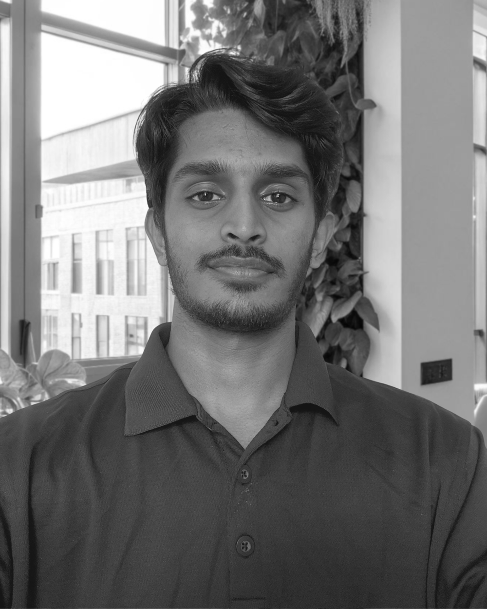 A young man with dark hair and a beard, wearing a dark collared shirt, taking a selfie indoors near a large window with a building and plants in the background.