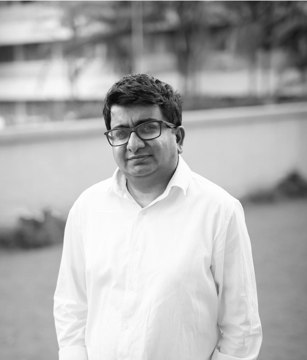 Black and white photo of a man with glasses wearing a white shirt, standing outdoors with a blurred background of trees and building.