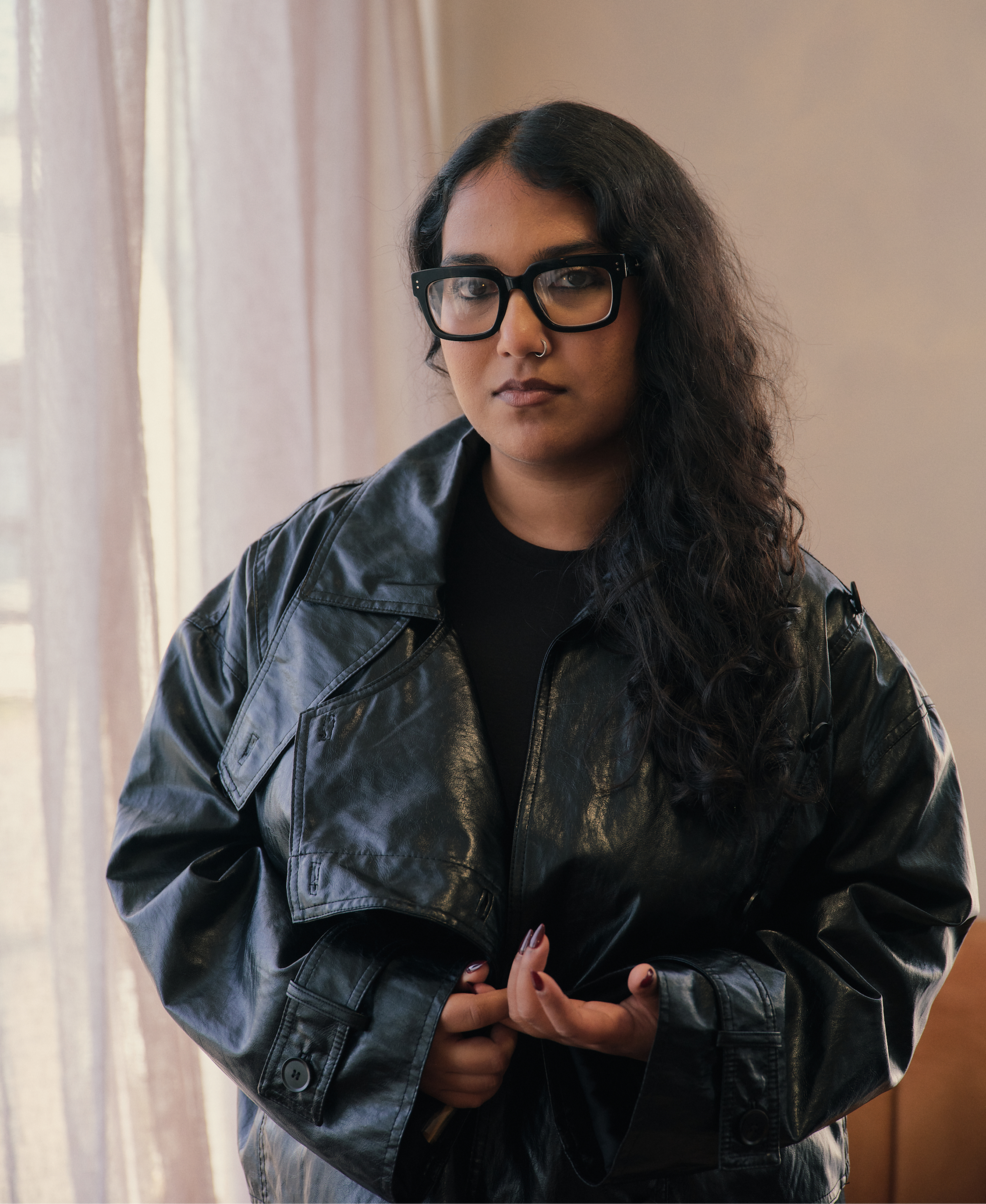 A woman with long dark curly hair, wearing large black glasses, a nose piercing, a black t-shirt, and a black leather jacket, standing indoors near a window with beige curtains.