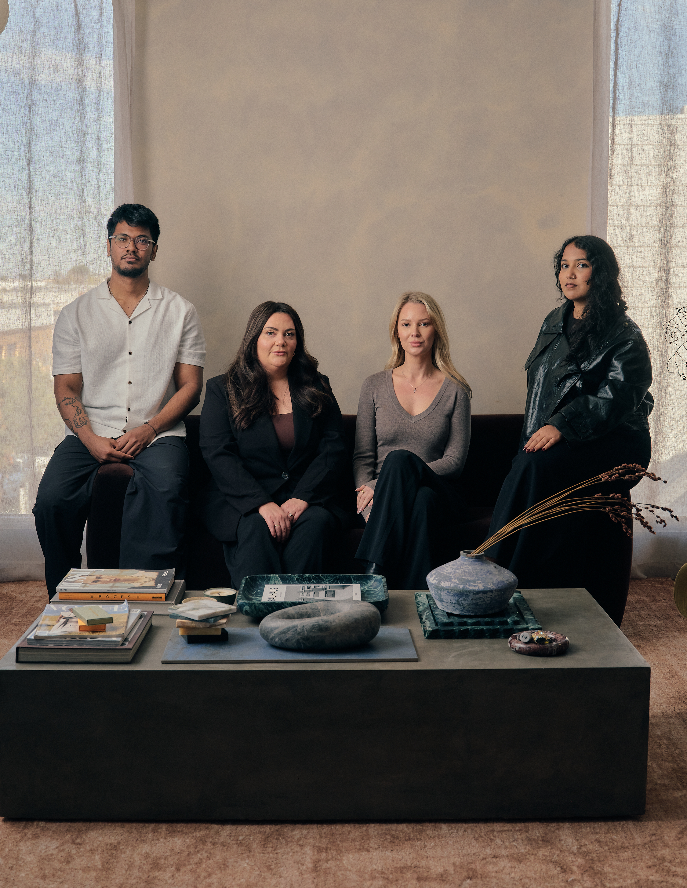 A group of four diverse women sitting and standing around a coffee table with books, magazines, and decorative objects in a well-lit room with curtains.