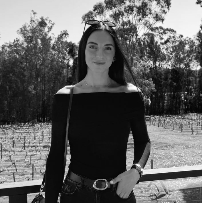 A woman with long dark hair wearing off-shoulder top, sunglasses on her head, and jewelry, standing outdoors with trees and vineyard in the background.