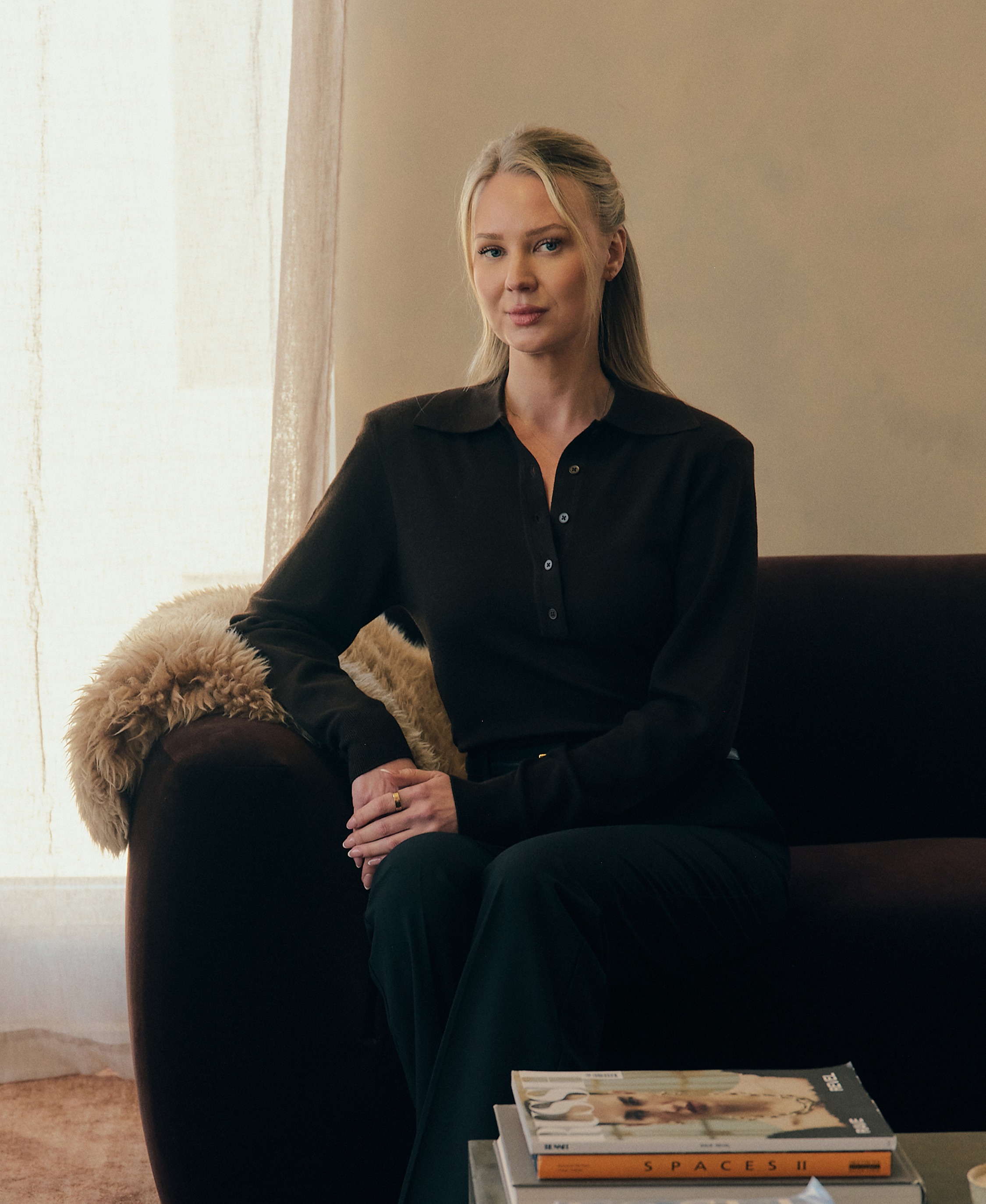 A woman with blonde hair and blue eyes sitting on a black chair in front of a beige wall with a window. Olivia Alexandersson owner and lead consultant of OLX STUDIOS Boutique Marketing Agency
