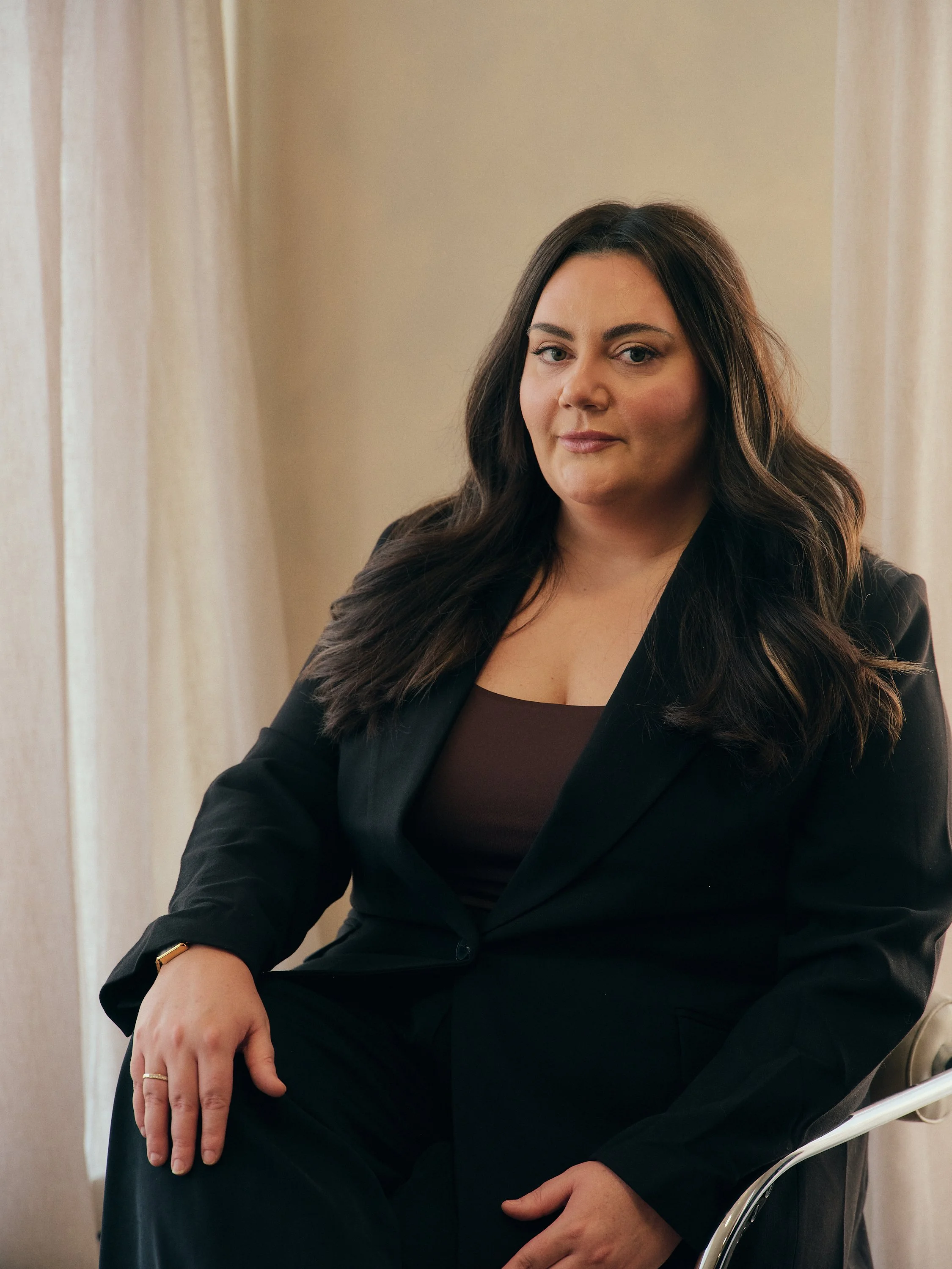 A woman with long dark hair, wearing a black blazer and brown top, sitting on a chair near cream-colored curtains.