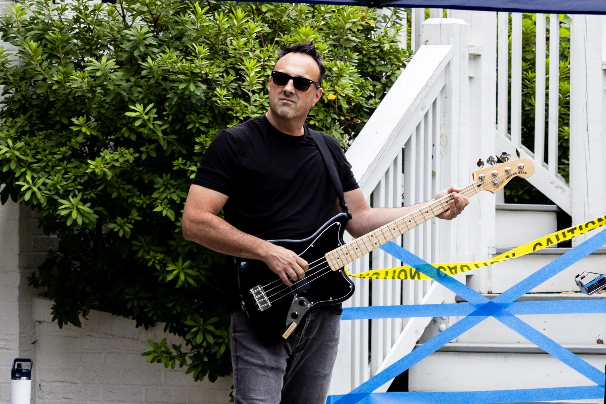 A man wearing sunglasses and a black t-shirt playing a bass guitar outdoors behind a white fence and green bushes, with yellow caution tape in front.