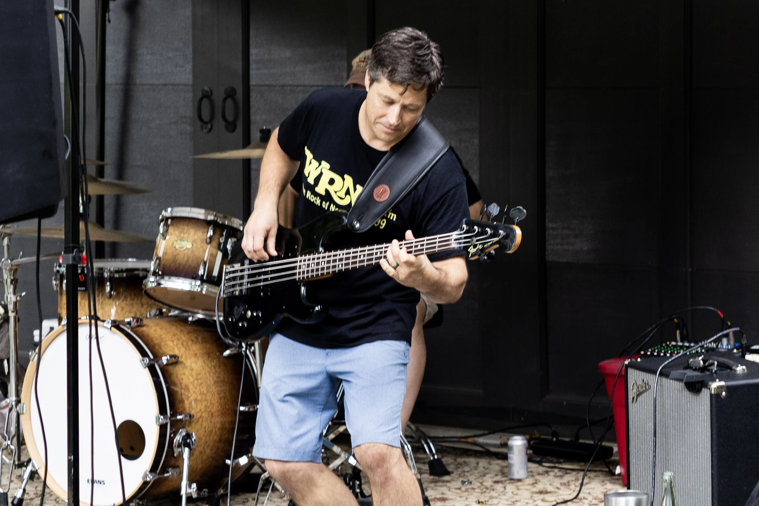 A man wearing a black t-shirt and light blue shorts playing an electric bass guitar during a live performance. There is musical equipment, including drums and amplifiers, visible in the background.