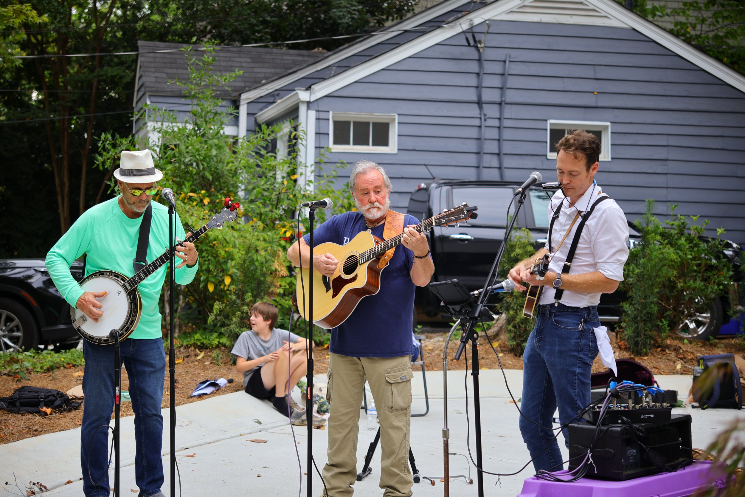 Three men playing musical instruments outdoors, with one playing a banjo, another a guitar, and the third a microphone, on a concrete surface in front of a house with trees and parked cars in the background.