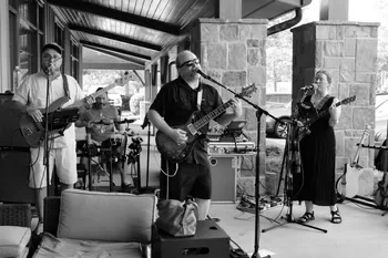 A group of five musicians performing outdoors under a covered patio, with stone pillars in the background. The band includes a man playing guitar and singing, a woman playing guitar and singing, a man playing drums, and a man playing bass guitar. There is a music stand and audio equipment on stage.