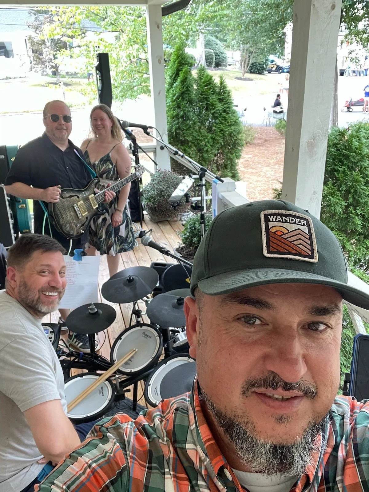 A group of four people, two men and two women, are on a porch with musical instruments; one man is taking a selfie with three others in the background, including a woman holding a guitar and another man playing an electronic drum set. They are smiling and appear to be enjoying a casual outdoor performance.
