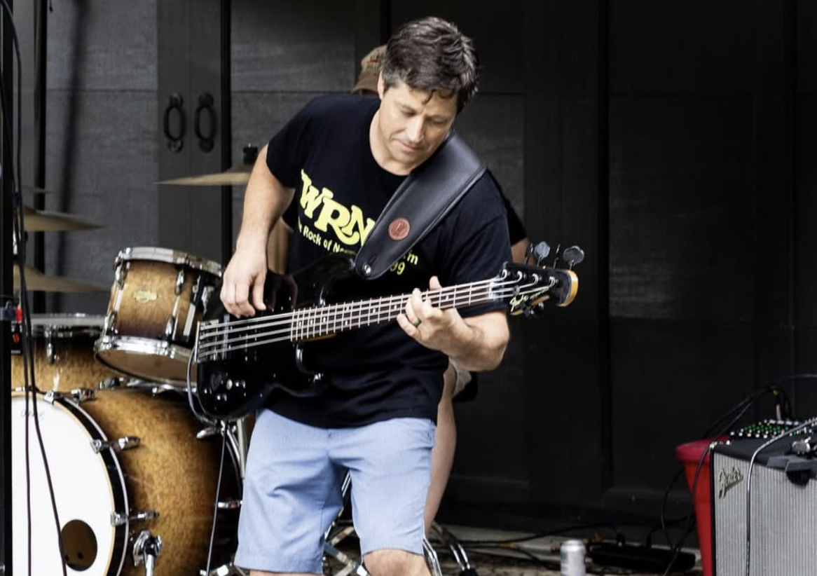 A man playing a black electric bass guitar during a live music performance, with drums and an amplifier visible in the background.