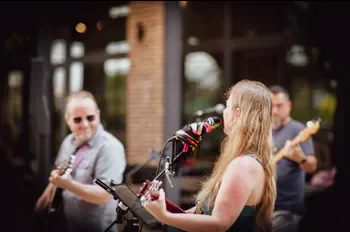 Group of musicians performing outdoors, with a woman singing into a microphone and playing guitar, and two men playing guitars in the background.