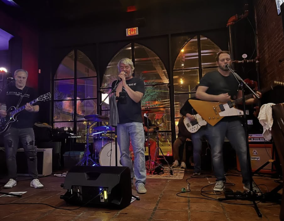 Band performing live music on stage in a dimly lit venue with brick walls and large windows. Four musicians playing guitars and singing into microphones.
