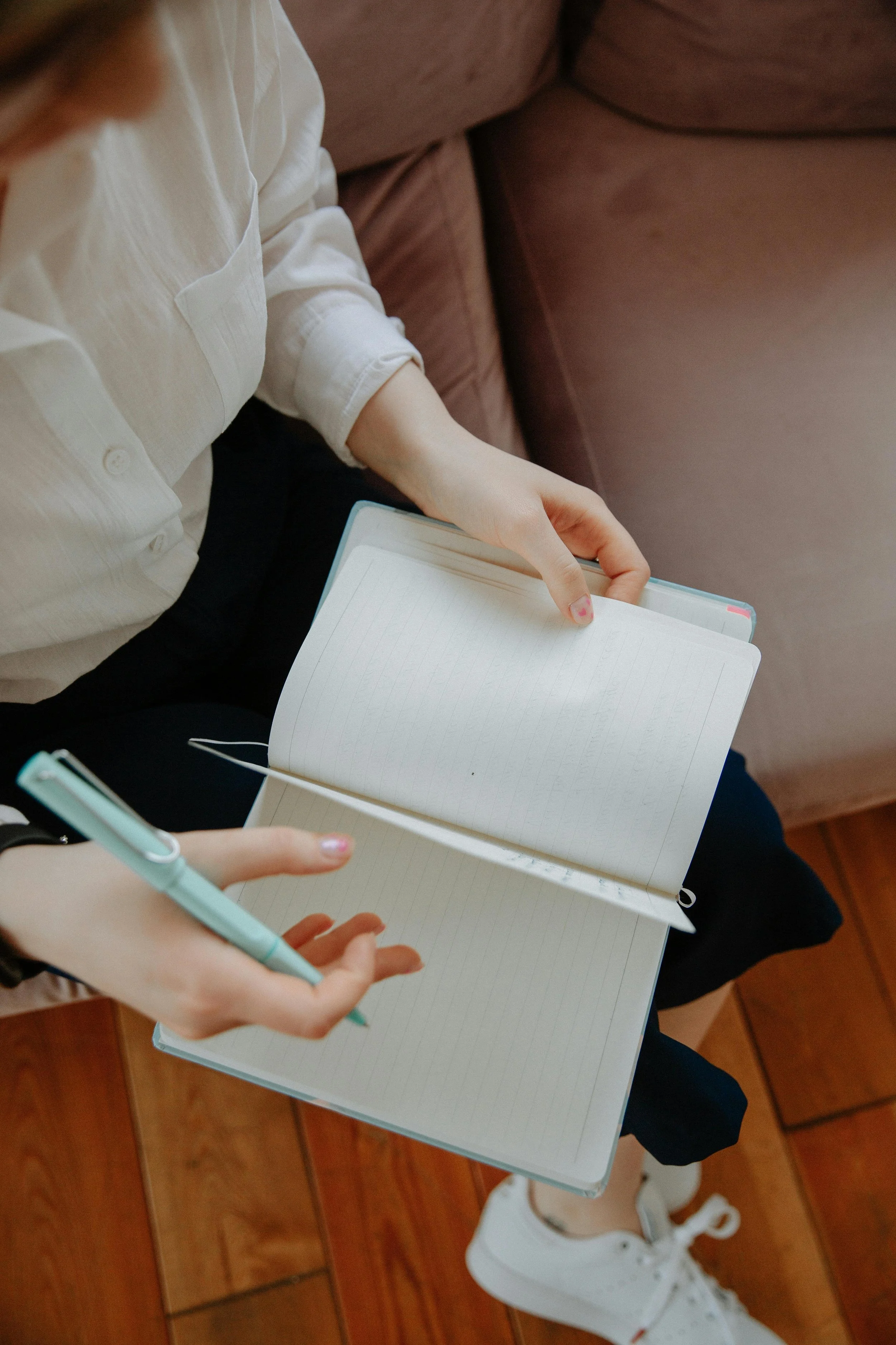 Two people sitting on a brown sofa, one holding a light green pen and a notebook, the other holding a notebook and pointing at it.