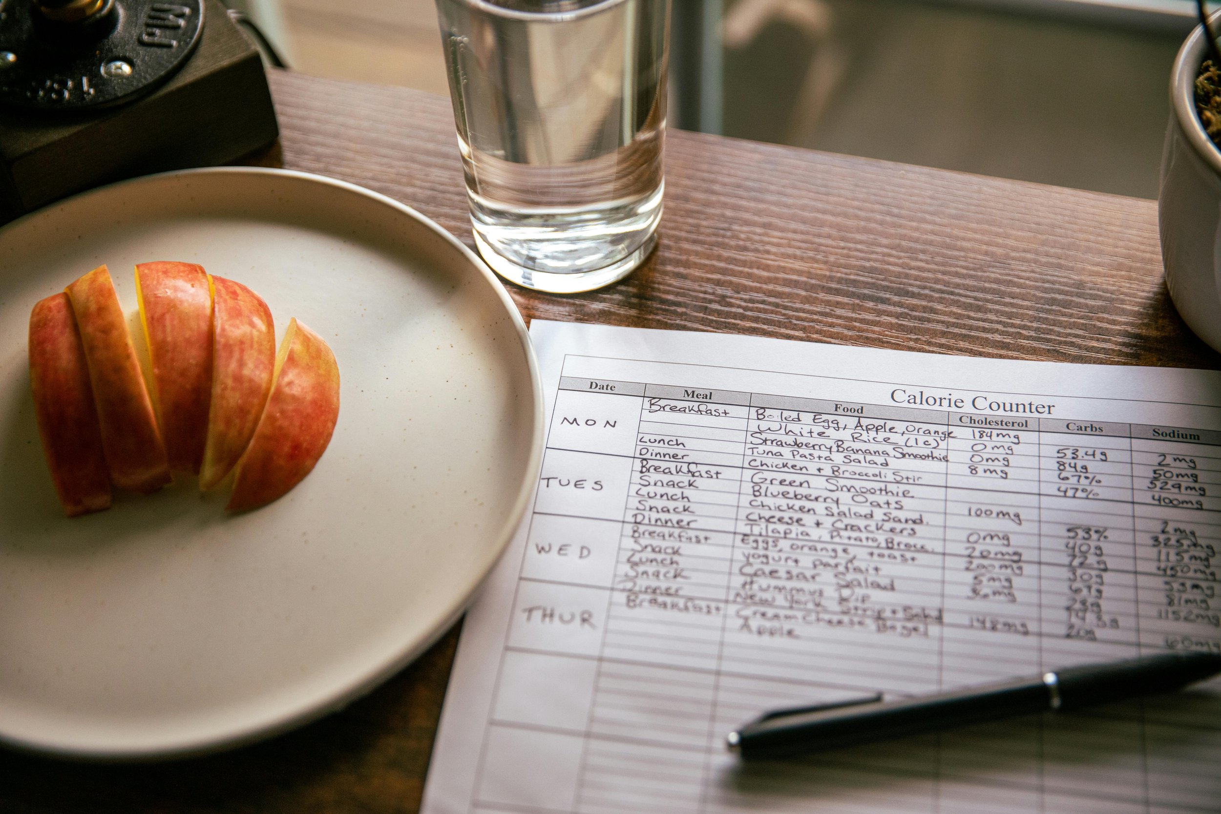 A wooden table with a glass of water, a black camera, a radio, a plate with sliced apple, and a handwritten calorie counter sheet showing meal plans and nutritional information.
