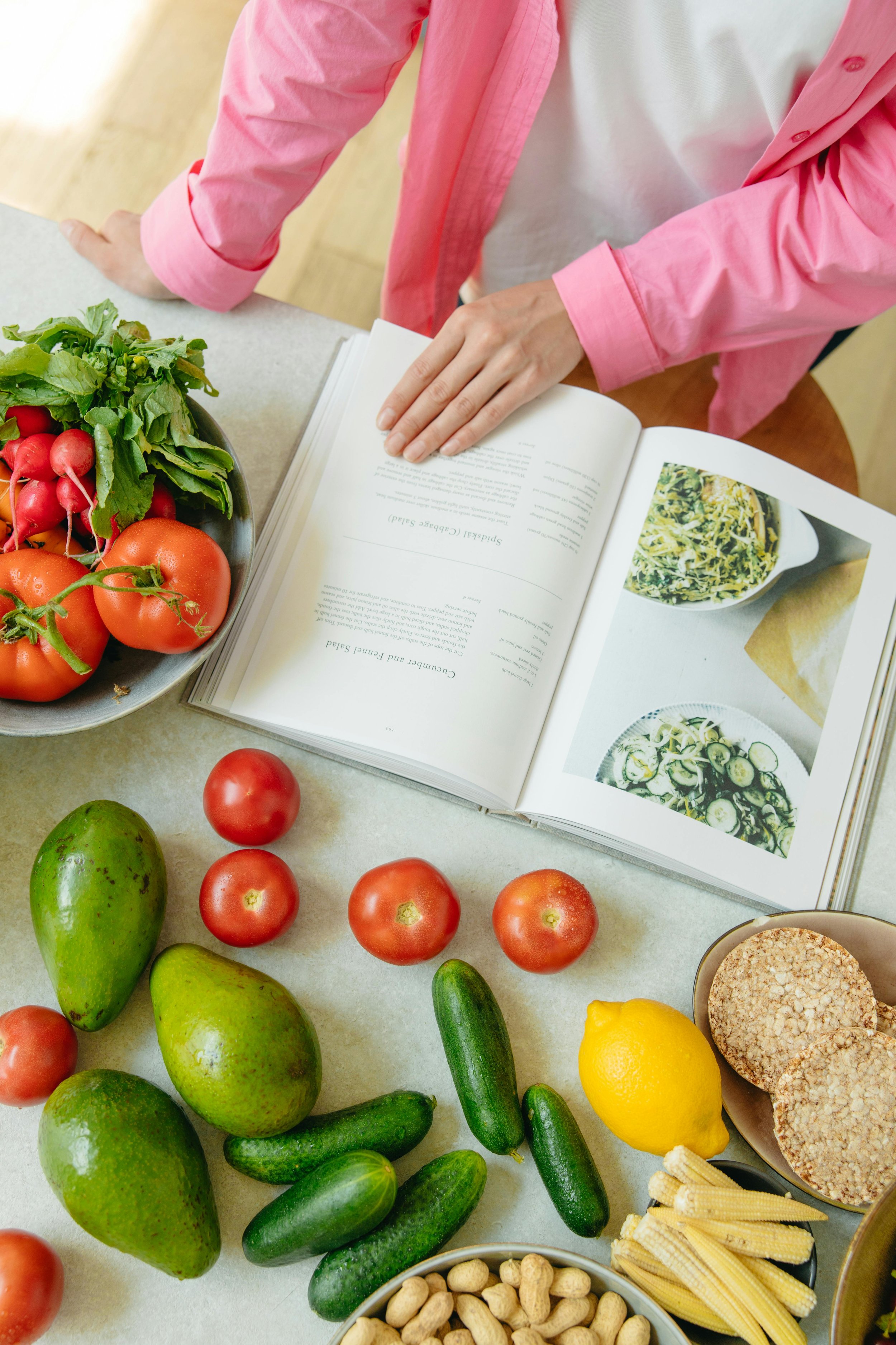 Fresh vegetables and ingredients on a kitchen counter, including tomatoes, radishes, cucumbers, avocados, lemons, baby corn, and a cookbook with recipes and images.