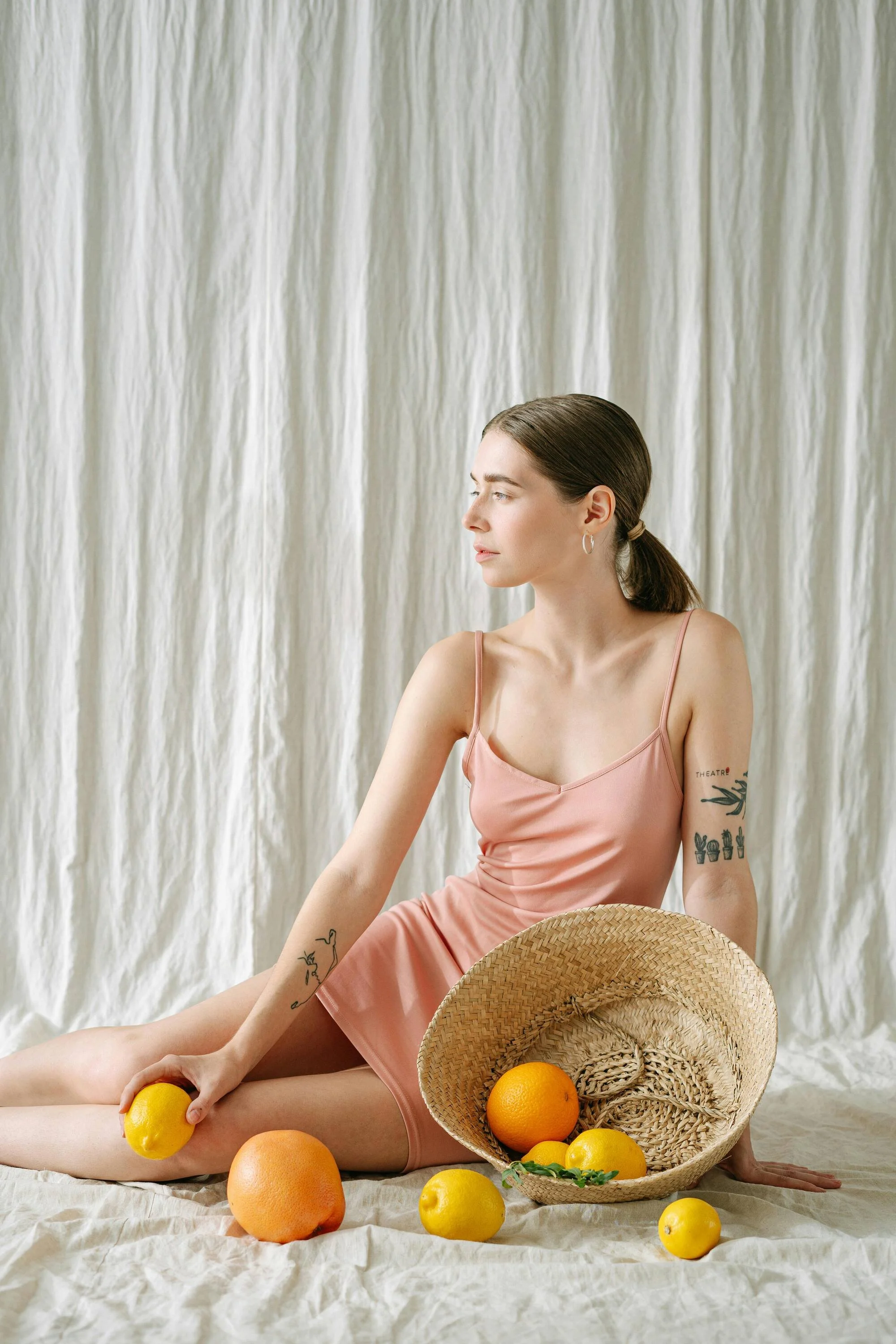 A woman sitting on a white fabric floor in front of a cream-colored curtain, holding a lemon, with various citrus fruits including oranges and lemons around her and a straw hat nearby.