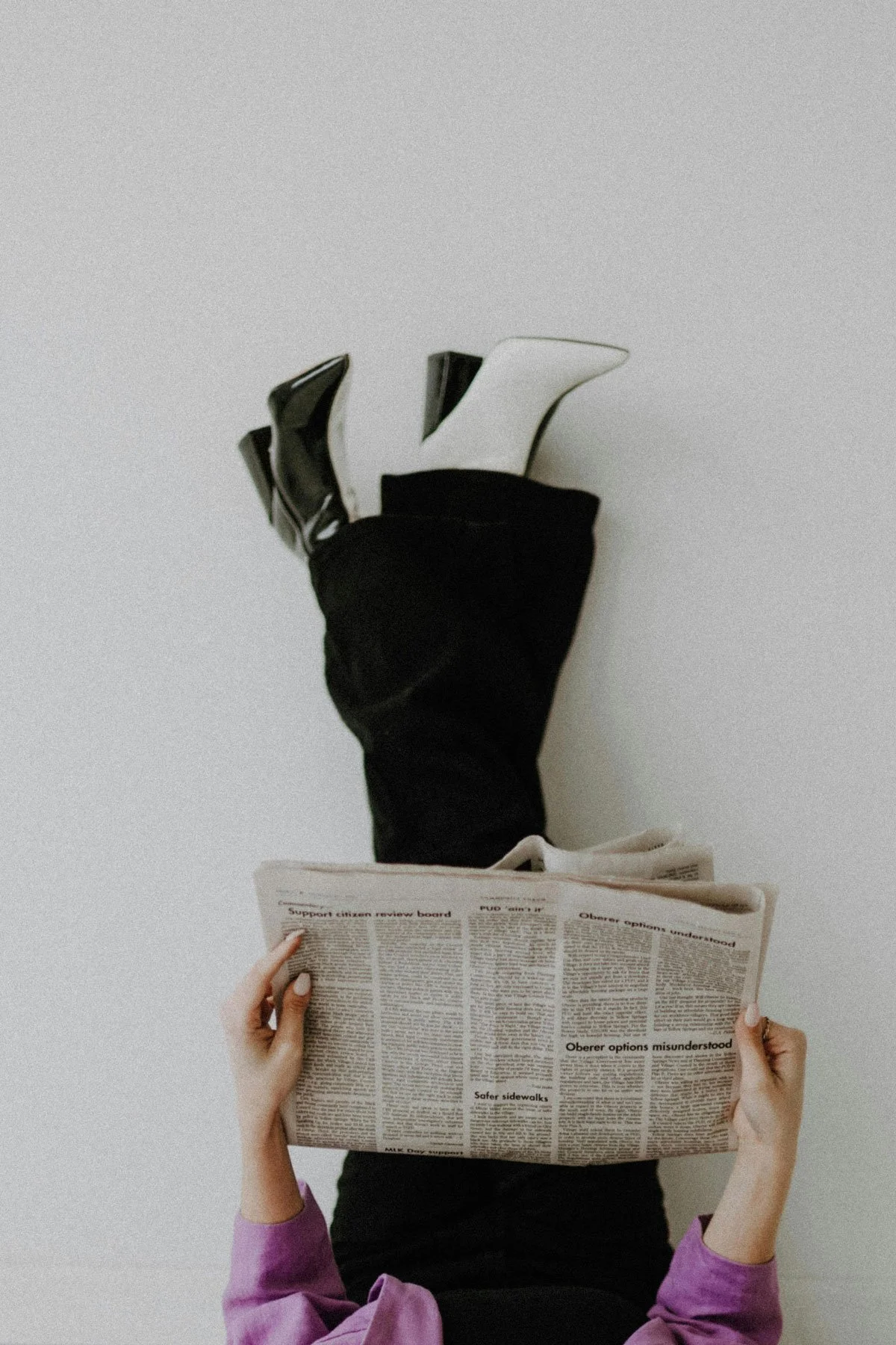 A person lying on the ground with their legs up against a wall, wearing black shoes and black pants, holding a newspaper. Another person's hands are visible holding the newspaper open, with arms in a purple long-sleeve shirt.