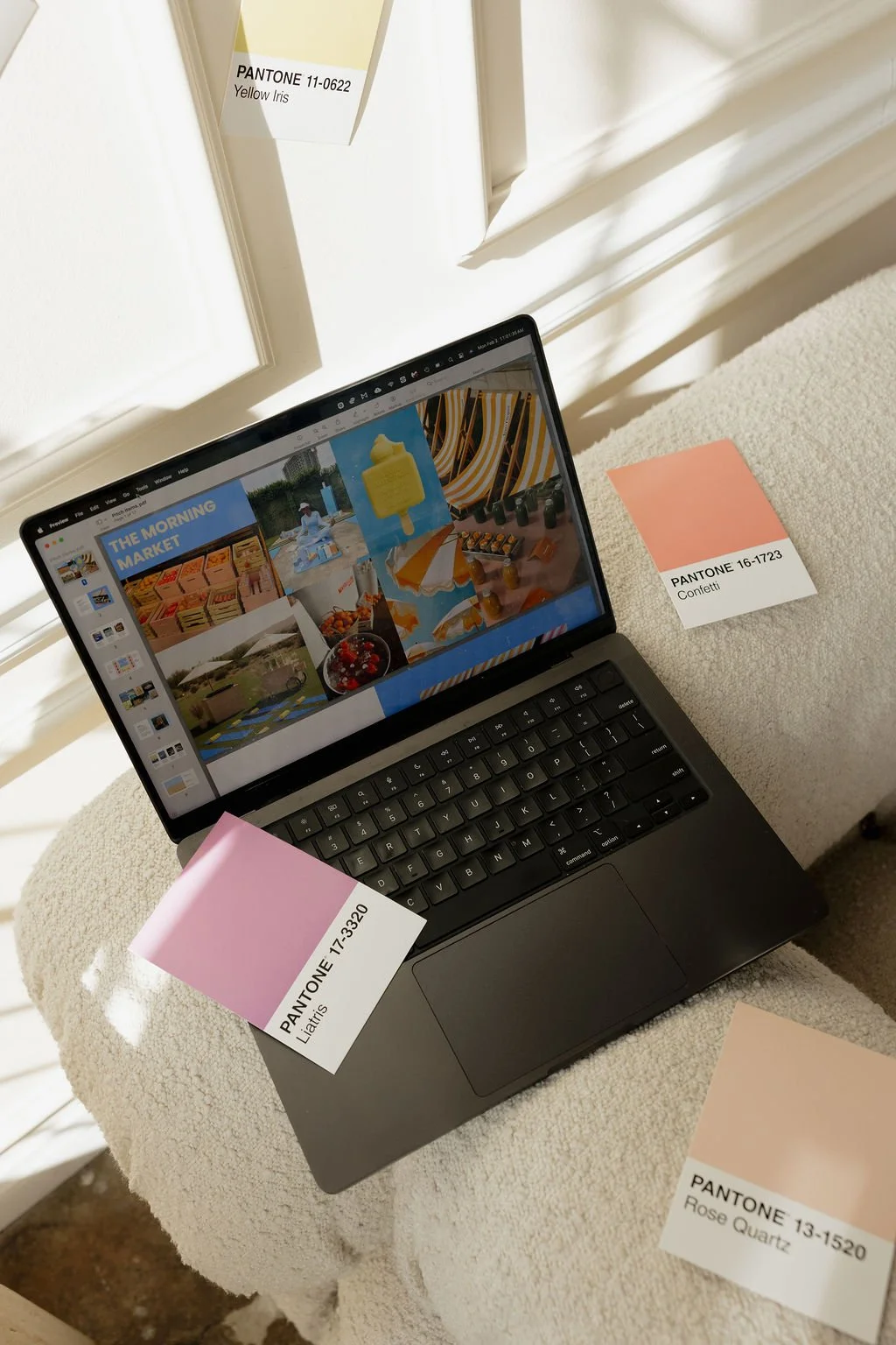 A laptop displaying a collage of outdoor summer scenes, with three color swatch cards labeled with Pantone colors and names on a beige textured surface. The cards are labeled Yellow Iris, Confetti, and Rose Quartz.