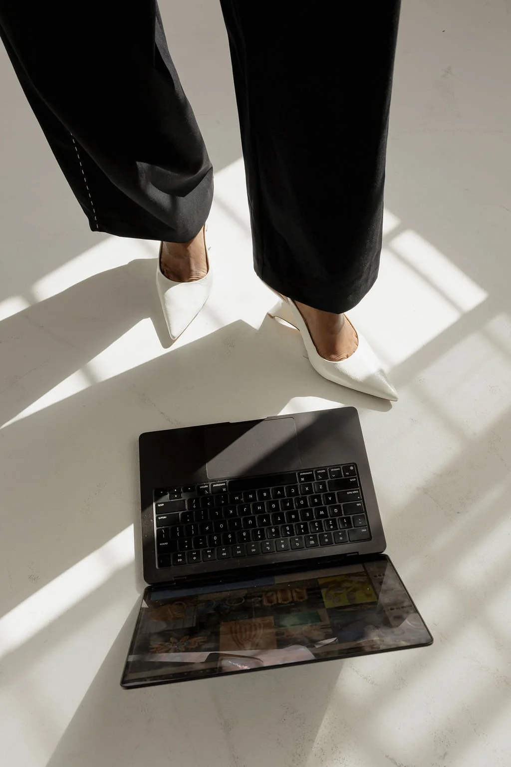 Person standing in black pants and white high-heeled shoes on a white floor with a black laptop open in front of them. Shadows from sunlight are cast on the floor.