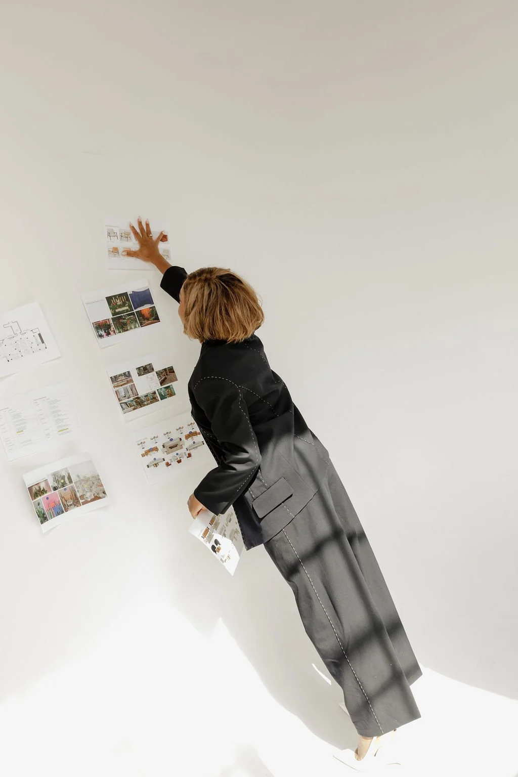 A woman in a black blazer and gray pants is standing and pointing at a white wall with various photographs and documents pinned on it.