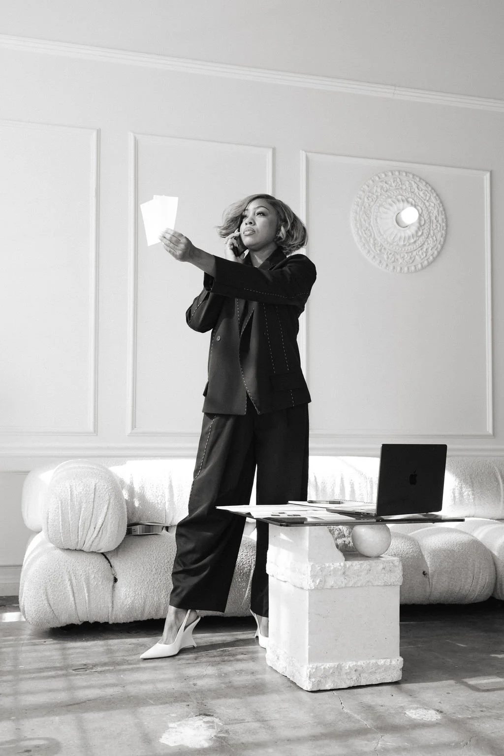 A woman in business attire stands in a modern, minimalist living room, talking on a phone, holding papers in one hand, with a laptop and magazines on a low table nearby.