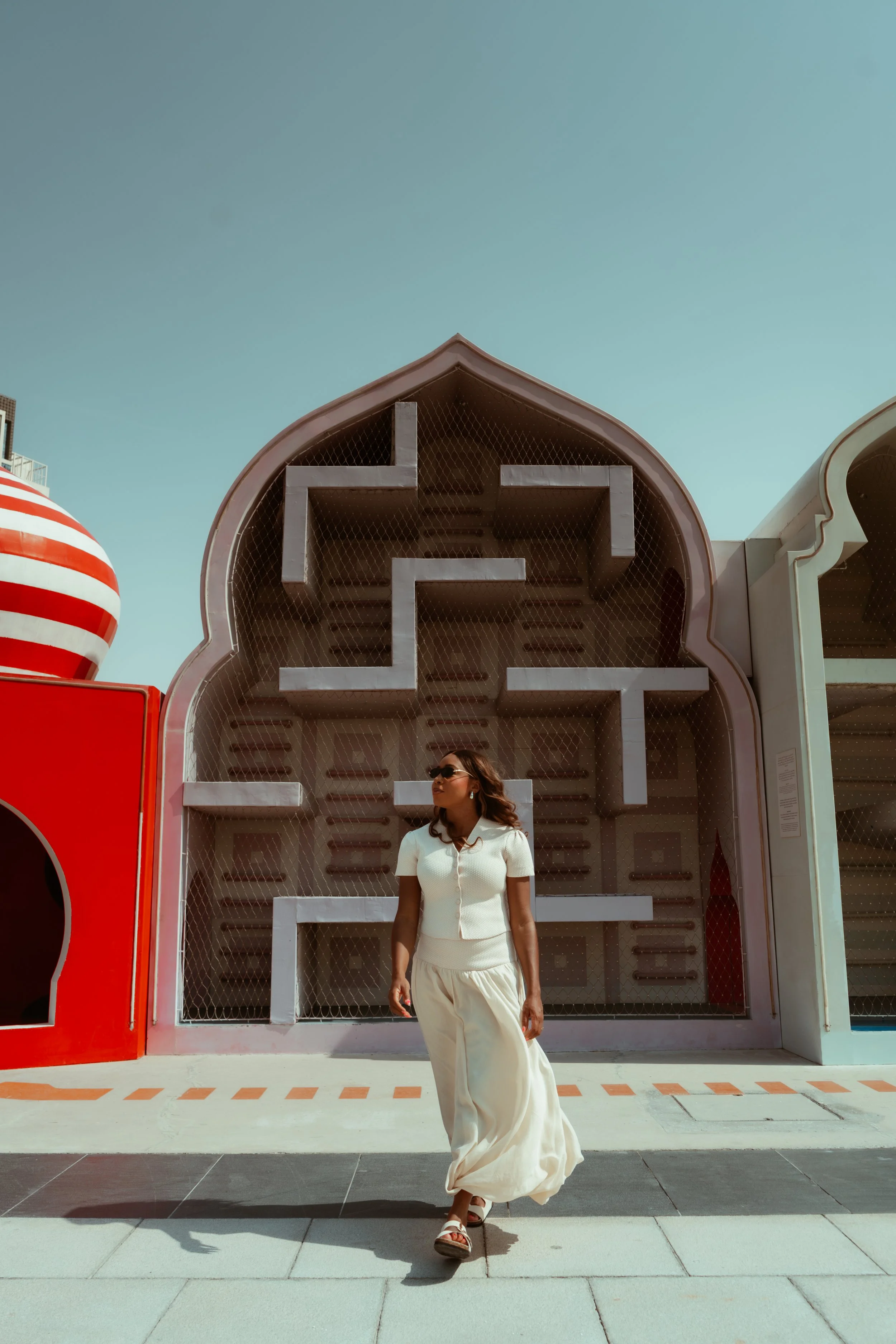 A woman wearing a white dress and sunglasses walking in front of a colorful, retro-style building with a large, red-and-white striped sphere and a geometric decorative structure.