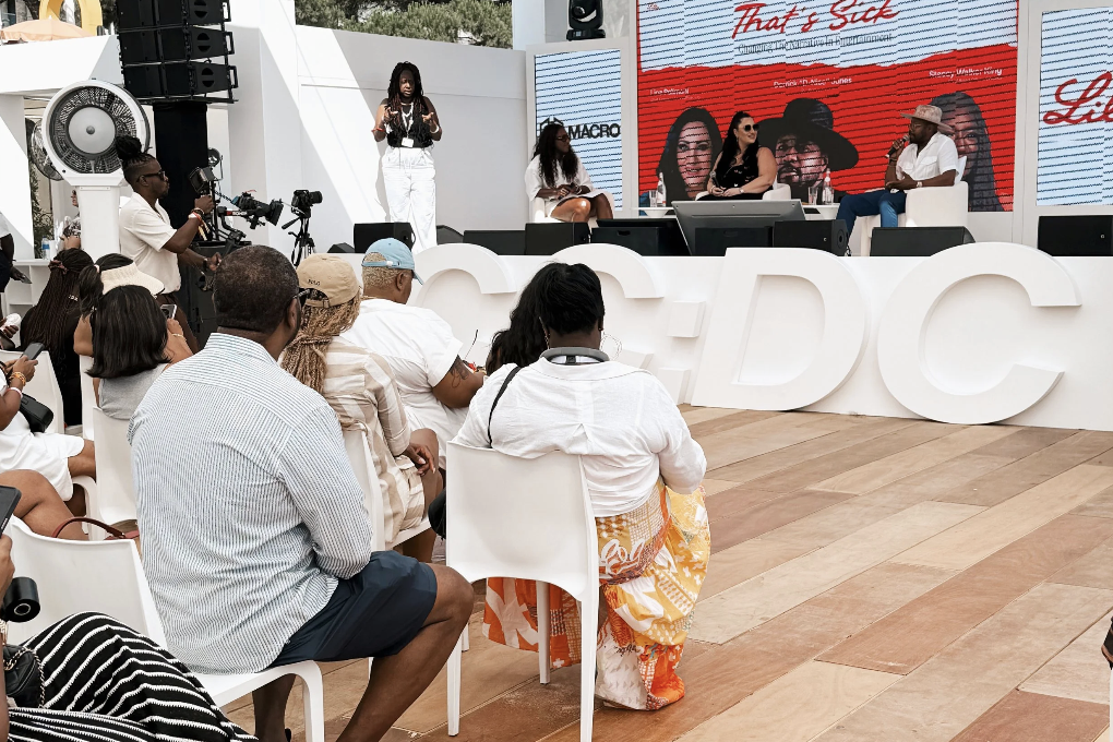 Panel discussion with four women on stage at Socially Equal event, audience seated, large white letters 'DC' in front, stage with screen behind displaying a red and white graphic.