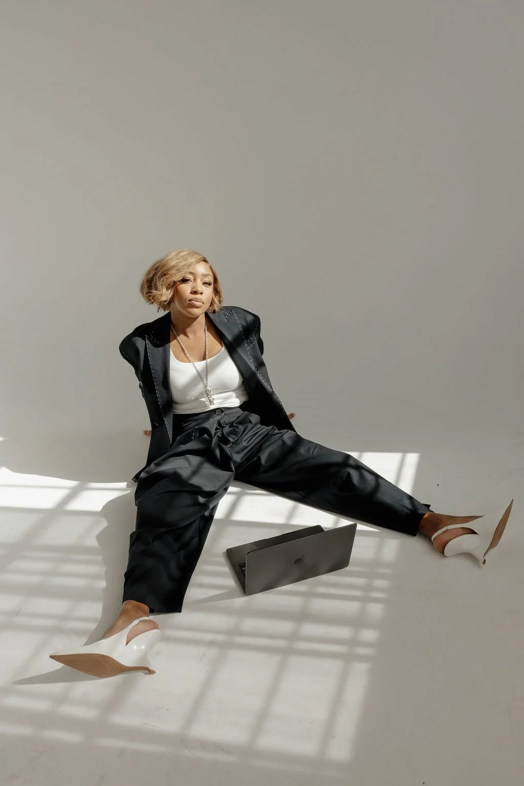 A woman with blonde hair sitting on a white floor, wearing a black jacket, white top, black pants, and white heels, with a closed laptop in front of her, and sunlight casting shadows through window blinds.