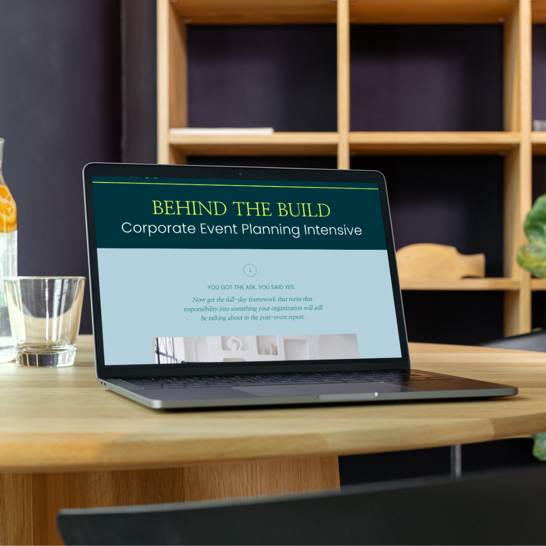 Laptop on wooden table displaying a presentation slide titled 'Behind the Build' related to corporate event planning, in a modern workspace with a glass and a plant nearby.